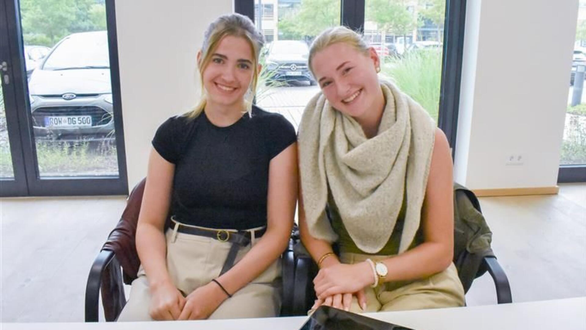 Zwei junge Frauen sitzen nebeneinander vor einem großen Fenster.
