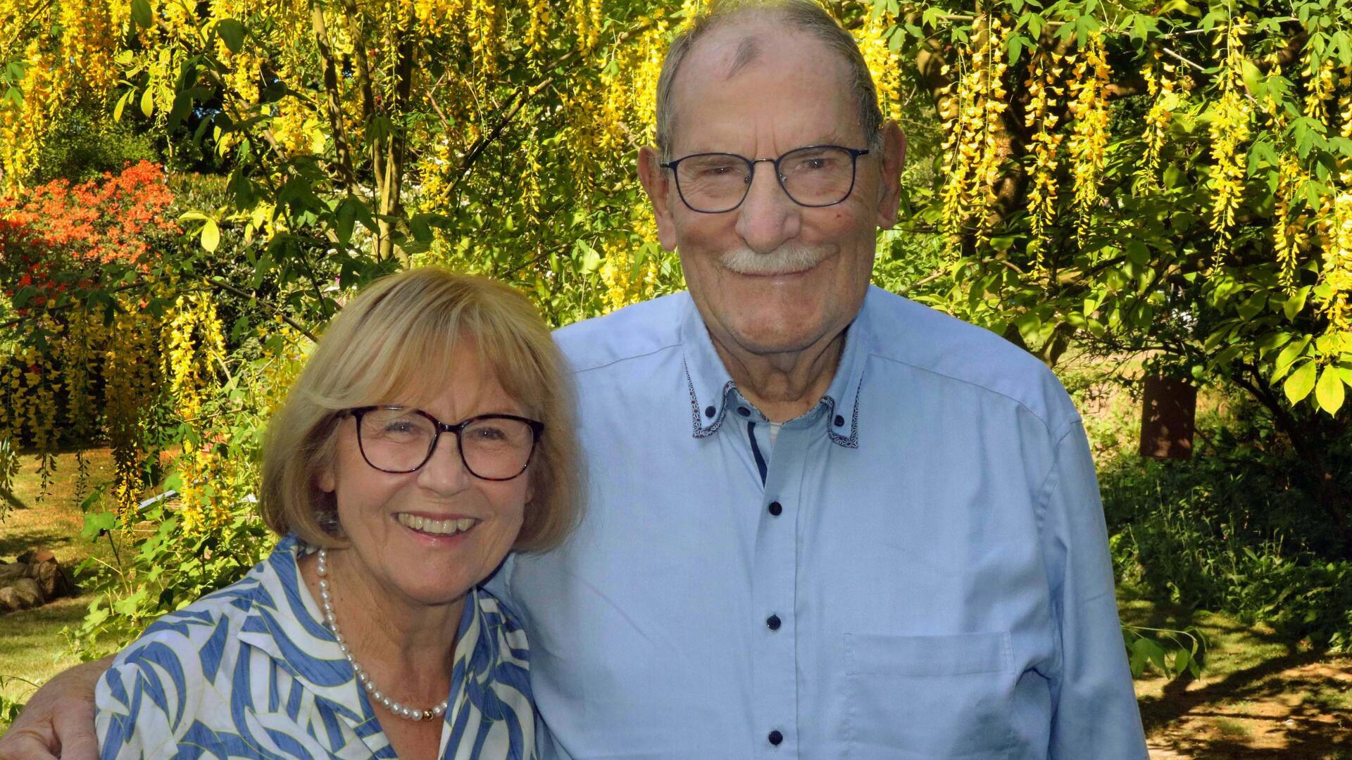 Edda und Hartmut Goede lächeln gemeinsam in die Kamera, umgeben von blühendem Garten mit gelben und roten Blüten.