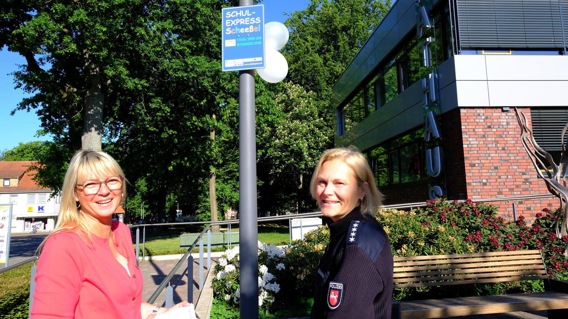 Zwei Frauen stehen neben einem Schild, auf dem „Schulexpress Scheeßel“ geschrieben steht. Eine von ihnen trägt eine Polizeiuniform.