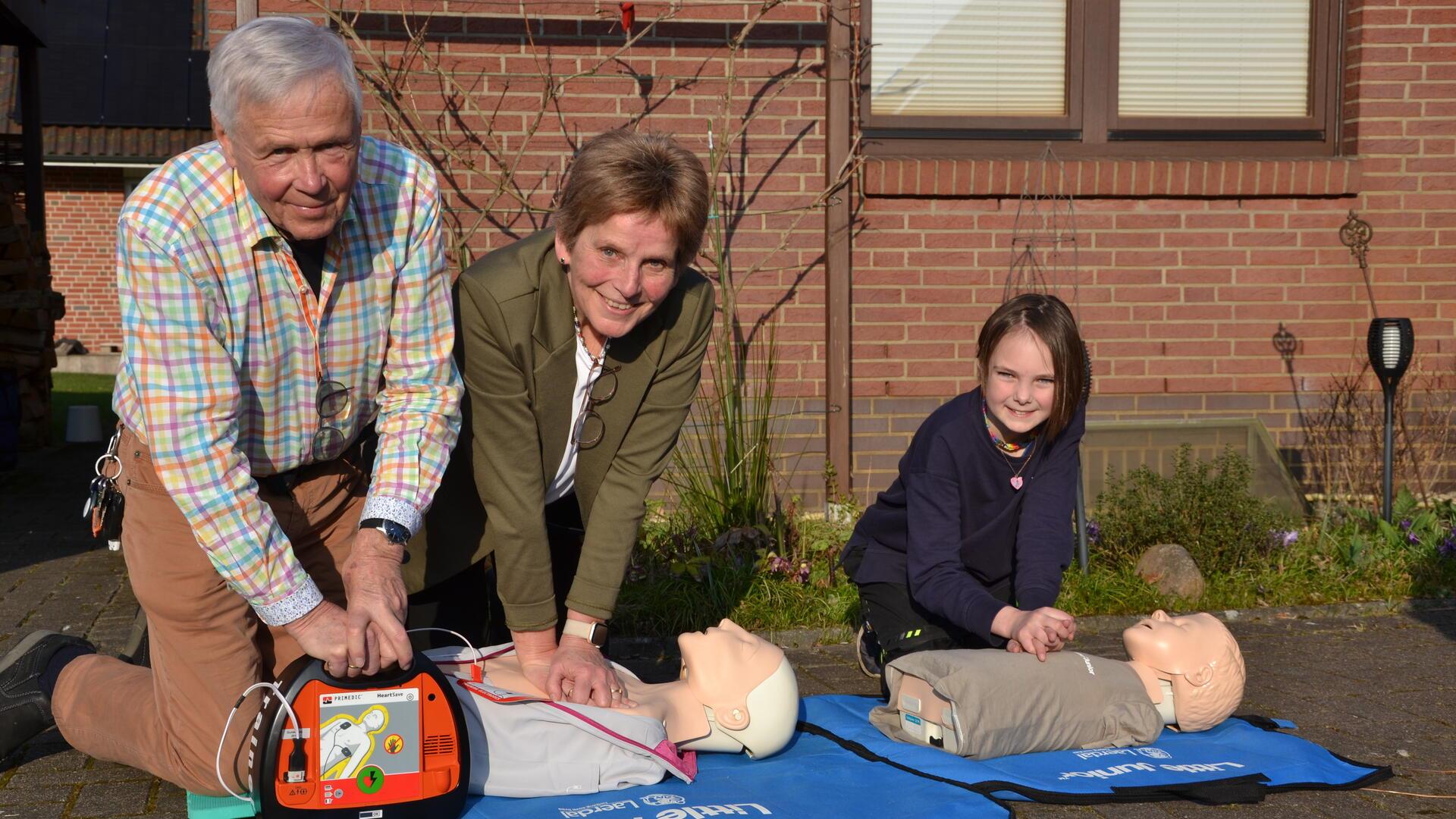 Ein Mann und eine Frau simulieren die Herzdruckmassage an einer Puppe, der Mann hält einen Defibrillator bereit. Neben ihnen simuliert ein Mädchen die Herzdruckmassage an einer Puppe.