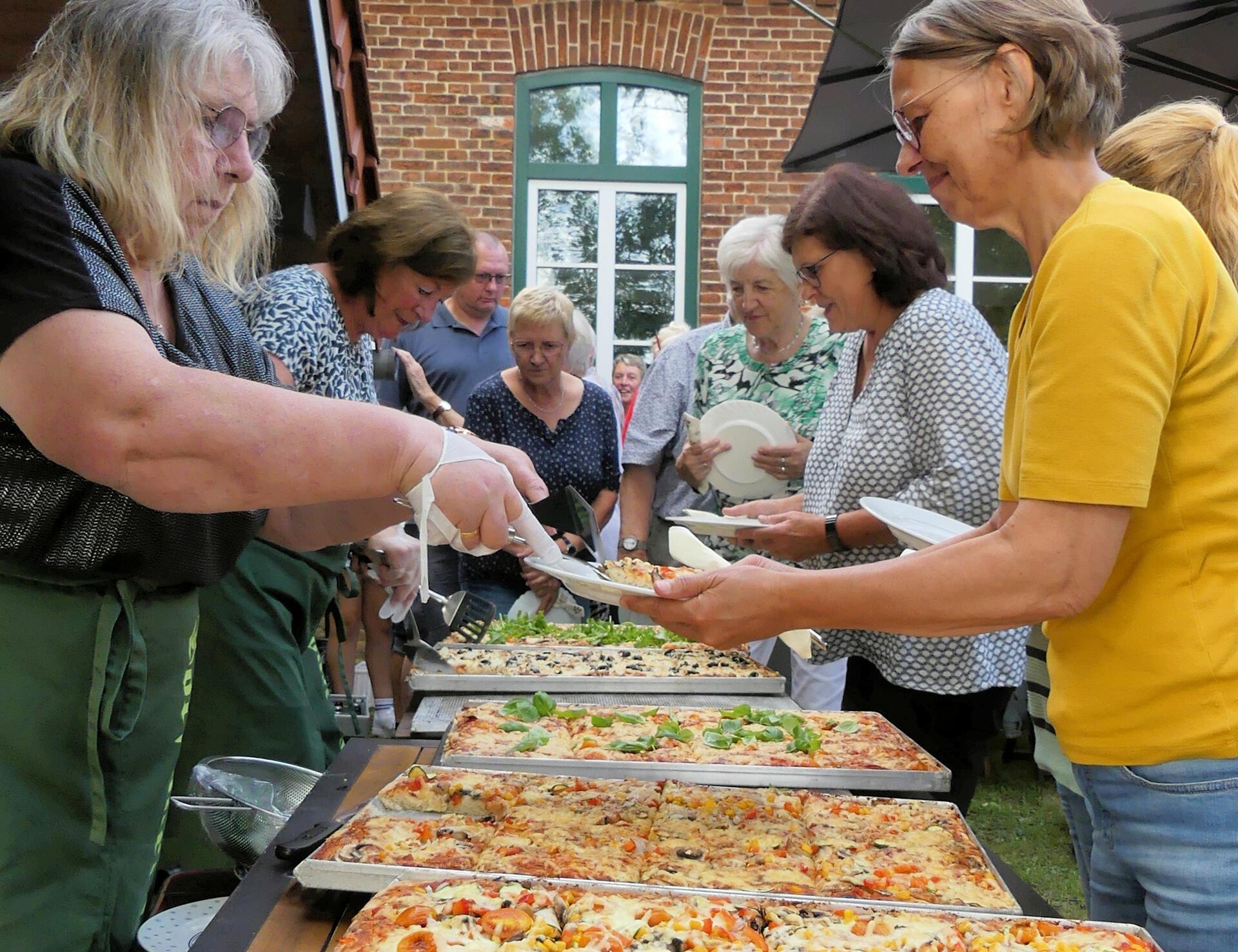 Eine Frau gibt Pizza von großen Backblechen aus. Andere Gäste warten mit Tellern in der Hand. 