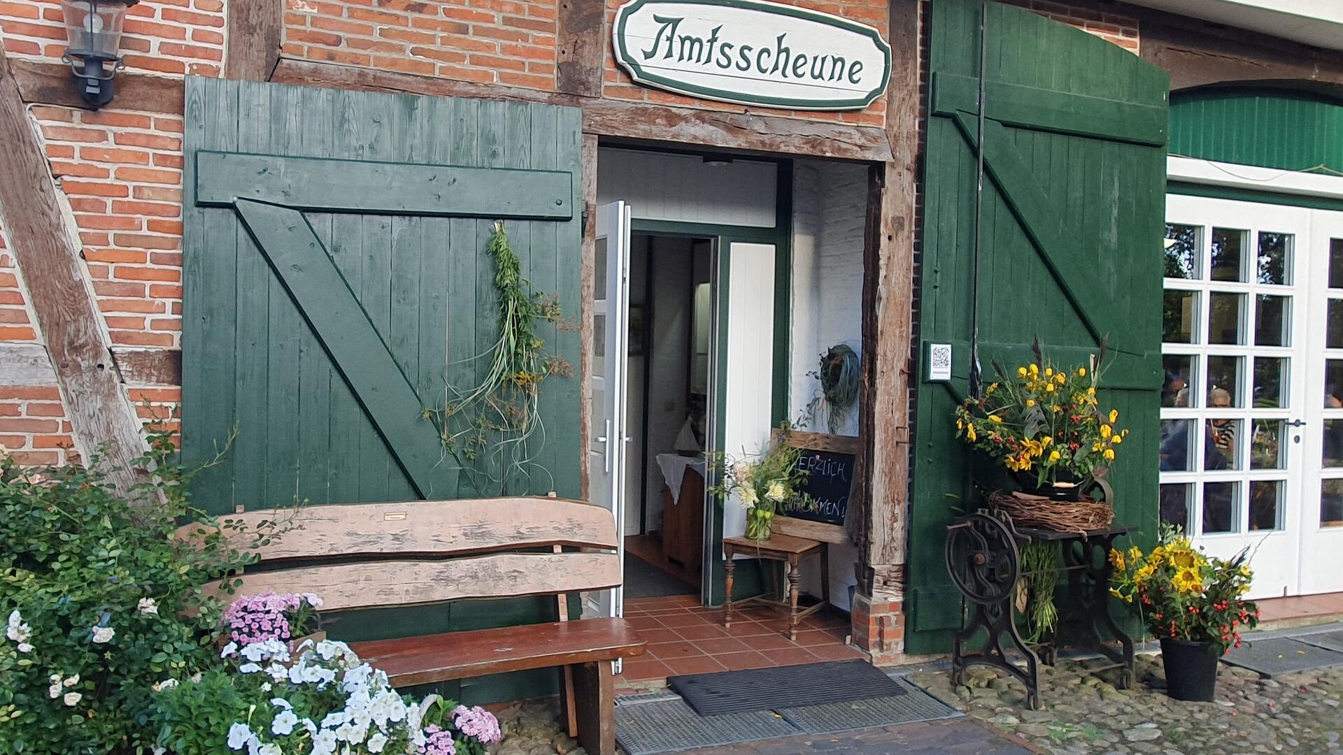 Eingang der Amtsscheune in Bad Bederkesa mit grün gestrichenen Holztoren, Blumenarrangements und einer Bank vor der Tür.