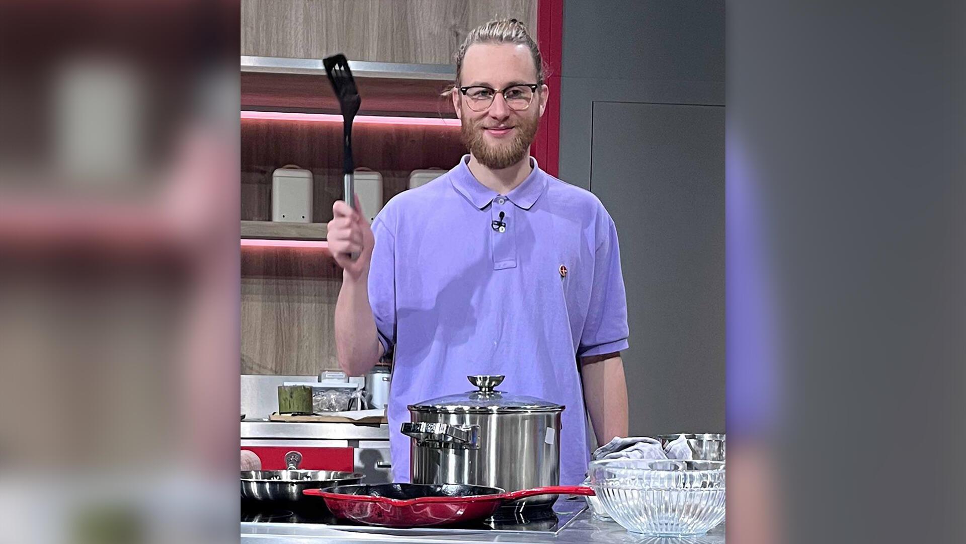 Ein junger Mann mit Brille und blondem Bart steht in einer modernen Küche. Er trägt ein lila Poloshirt mit einem kleinen Logo und hält eine schwarze Pfannenwender hoch. 