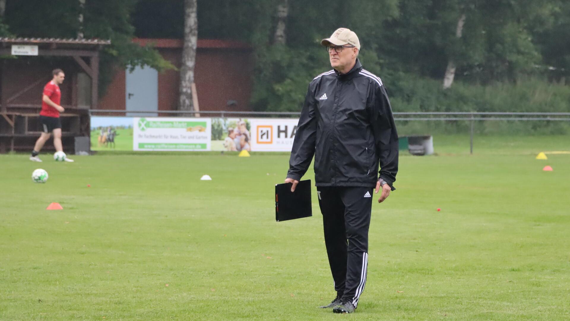 Ein Mann in einem schwarzen Sportdress und einer Cap hält eine Mappe in der Hand und geht über einen Rasen.