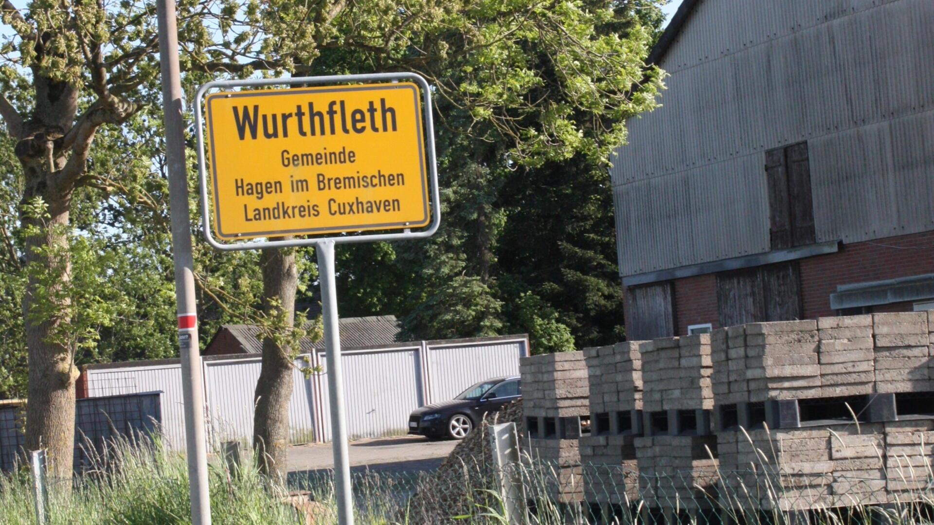 Ortsschild von Wurthfleth in der Gemeinde Hagen im Bremischen, Landkreis Cuxhaven, vor einer Halle mit gestapelten Betonplatten und umgeben von Bäumen.