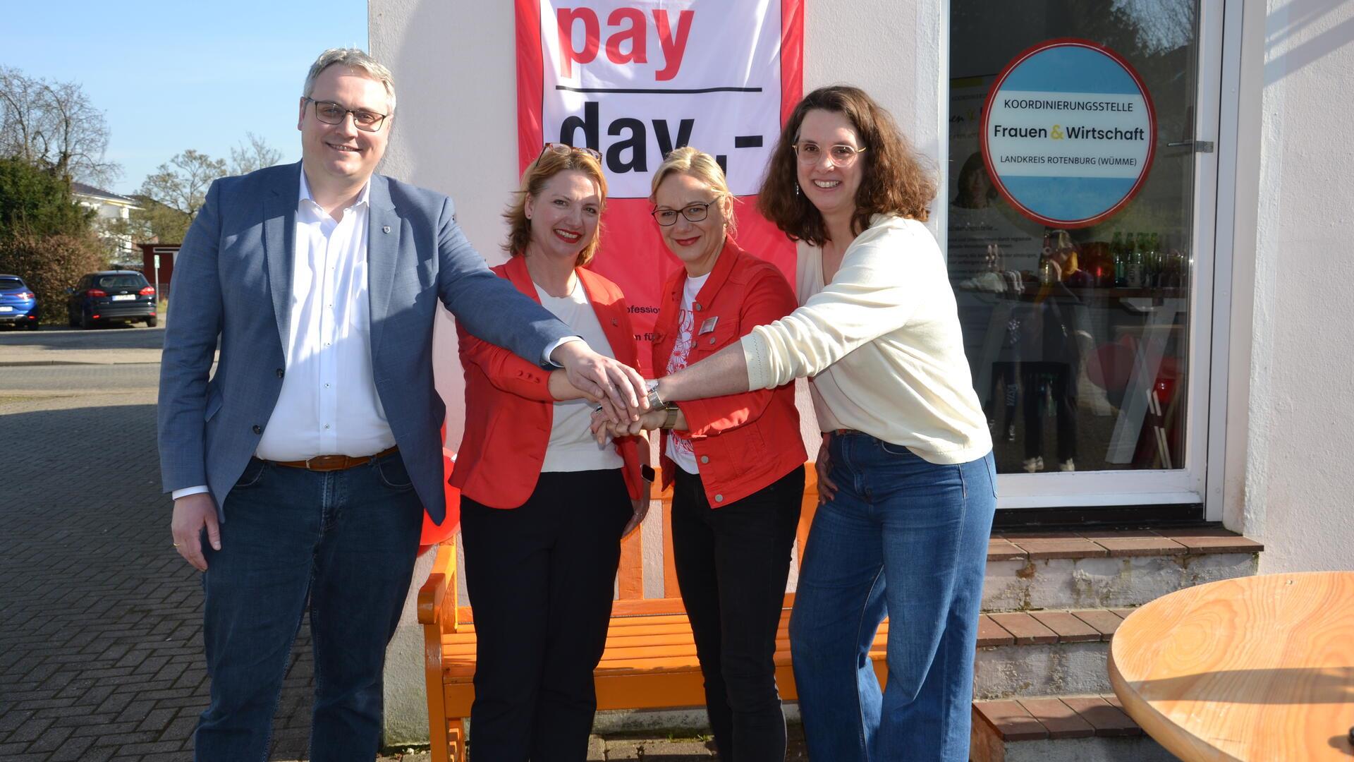 Ein Mann und drei Frauen stehen vor einem Plakat mit der Aufschrift "Equal Pay Day" und legen ihre Hände übereinander.