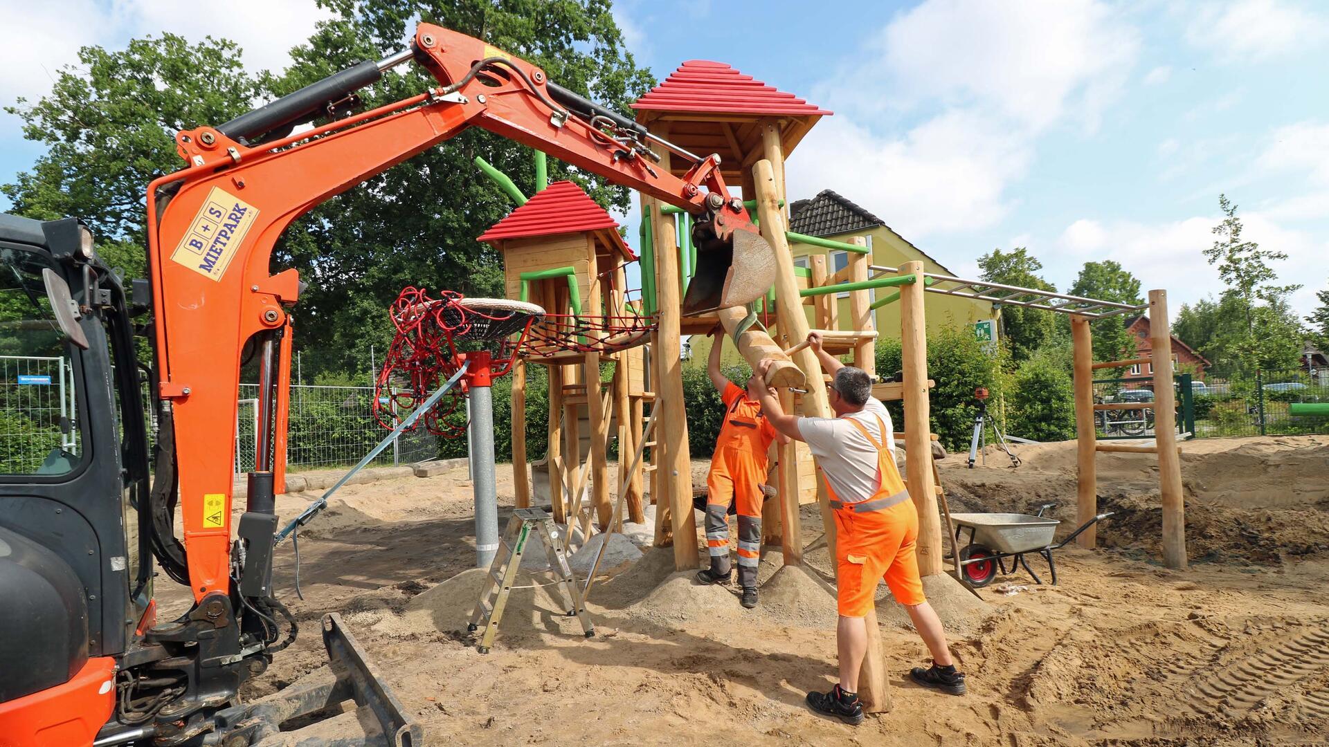 Zwei Bauarbeiter montieren mit Hilfe eines Baggers ein neues Holzspielgerät mit Rutsche auf einem sandigen Schulhofgelände. Im Hintergrund sind weitere Spielgerüste und ein Wohnhaus zu sehen.