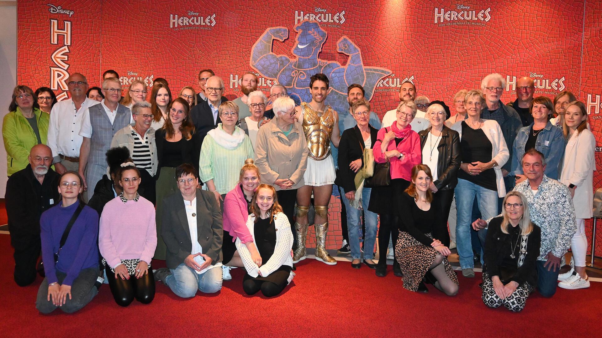 Rund 50 Menschen stehen als Gruppe beisammen. In der Mitte: ein Musicaldarsteller in Hercules-Kostüm.