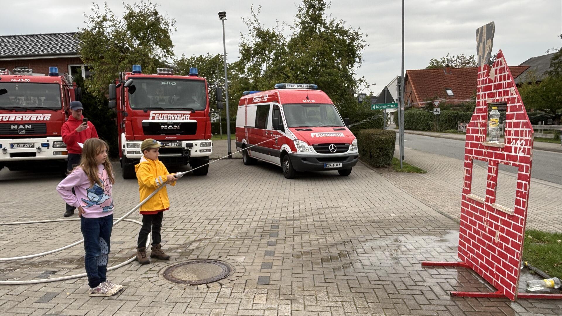 Zwei Kinder üben mit einem Feuerwehrschlauch das Zielspritzen auf ein stilisiertes Hausmodell, im Hintergrund stehen mehrere Feuerwehrfahrzeuge.