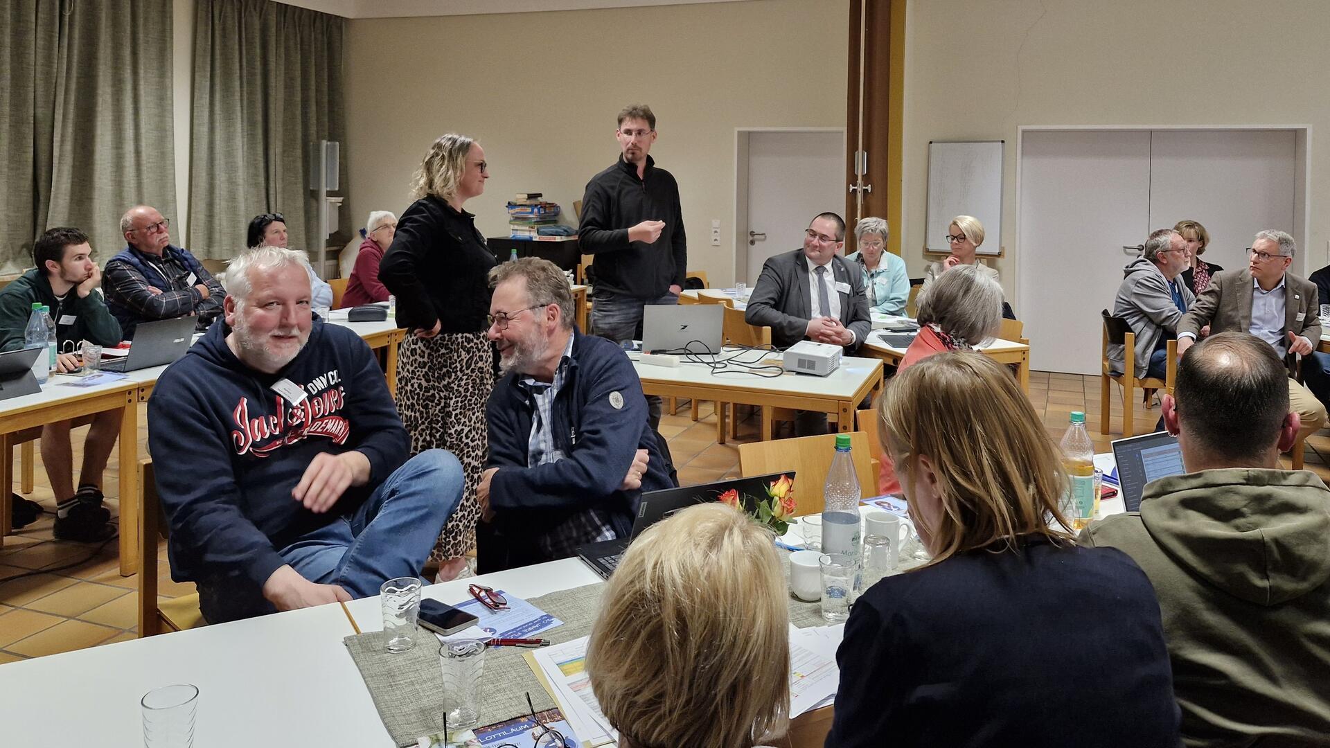 Menschen sitzen in Gruppen an Tischen in einem Konferenzraum und diskutieren lebhaft während einer Sitzung. Einige Teilnehmende arbeiten an Laptops, andere stehen oder unterhalten sich. Auf den Tischen liegen Getränke, Unterlagen und Namensschilder.