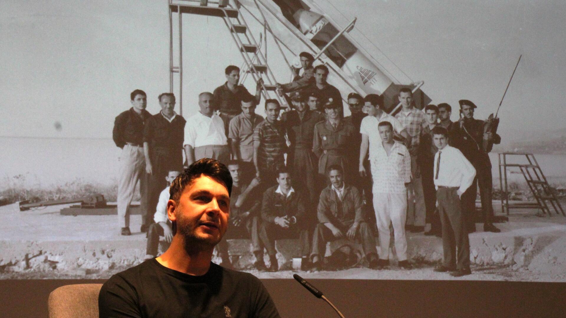Ein Mann sitzt vor einem Mikrofon auf einer Bühne, hinter ihm ist ein historisches Schwarz-Weiß-Foto projiziert, das eine Gruppe von Männern vor einer Rakete zeigt.