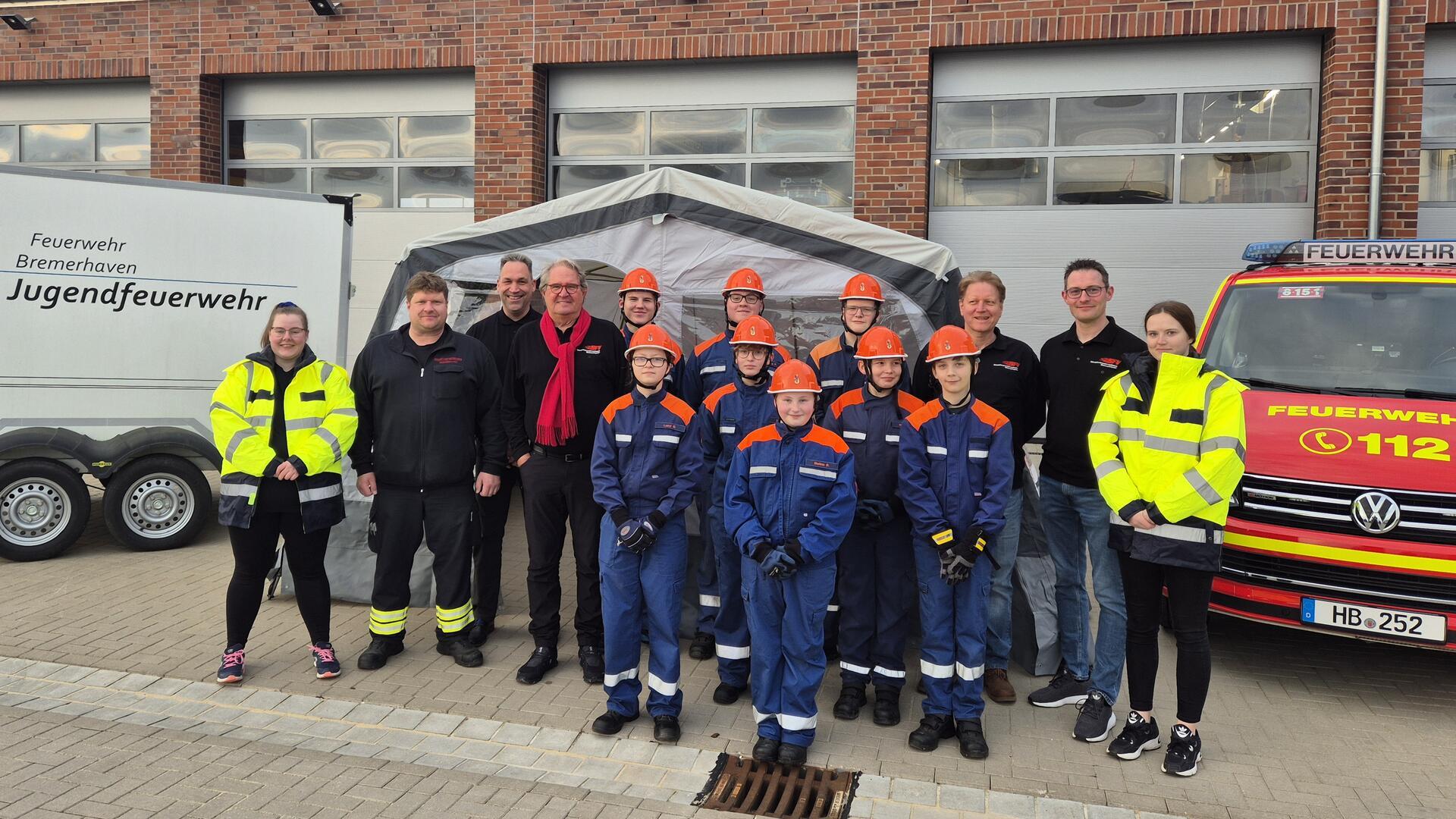 Gruppenfoto vor einer Feuerwehrwache mit Mitgliedern der Jugendfeuerwehr Bremerhaven und Betreuenden. Im Hintergrund sind ein neues Zelt, ein Feuerwehranhänger mit der Aufschrift „Jugendfeuerwehr“ sowie ein Einsatzfahrzeug zu sehen. Die Jugendlichen tragen blaue Schutzanzüge mit orangefarbenen Helmen.