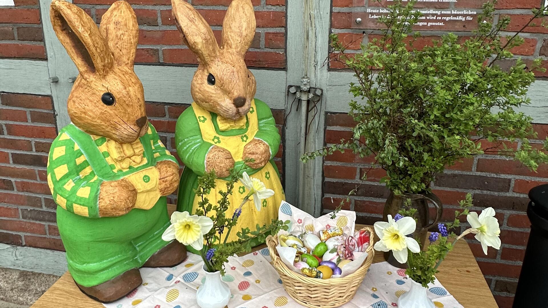 Zwei große Osterhasenfiguren stehen auf einem Tisch neben Blumen und einem mit bunten Eiern gefüllten Osternest. 