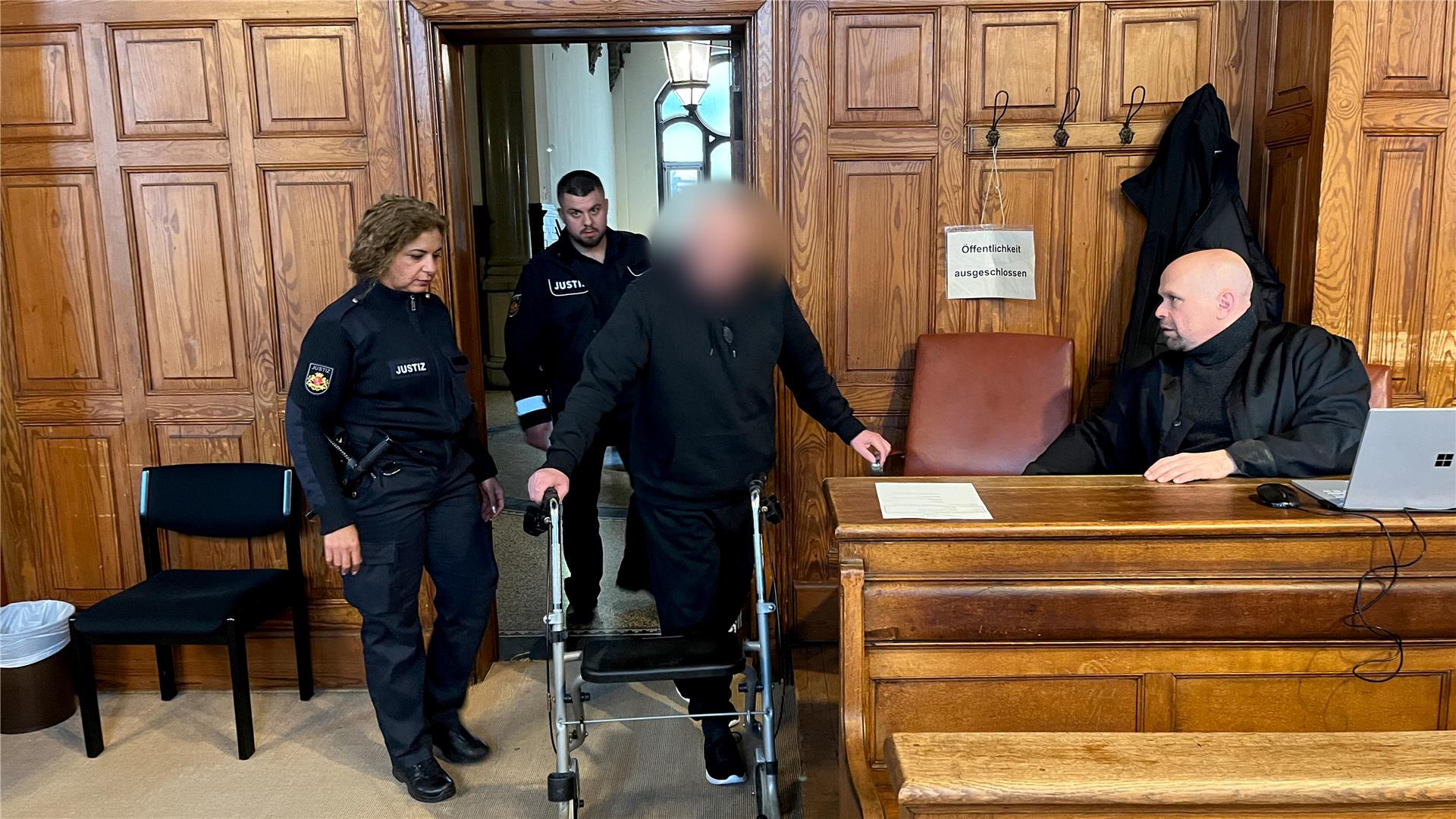 Angeklagter wird am Rollator von Justizbeamten zur Anklagebank geführt. 