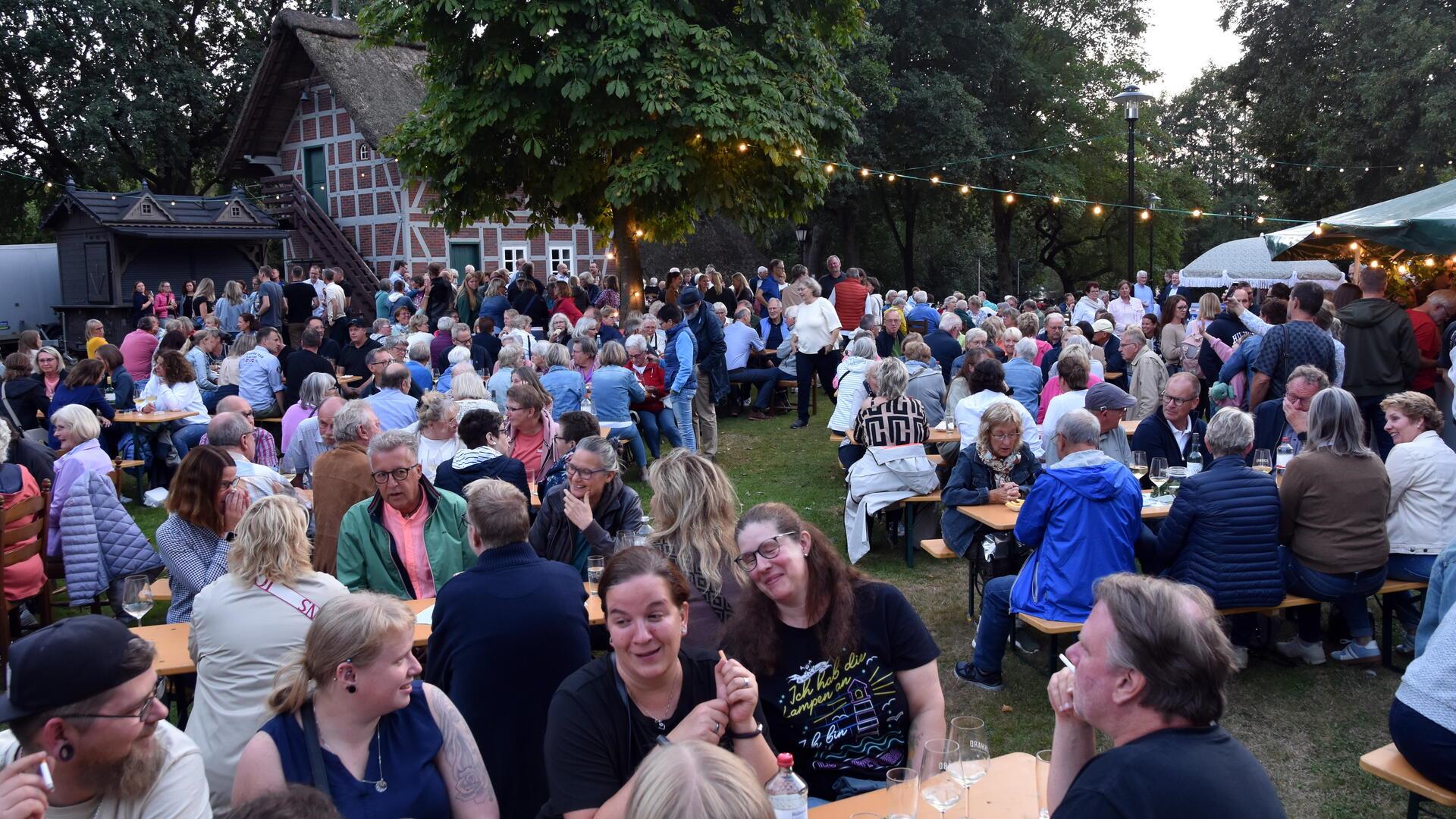 Unzählig viele Menschen sitzen an Biertischgarnituren vor einem norddeutschen Fachwerkhaus, im Hintergrund stehen auch viele Menschen.