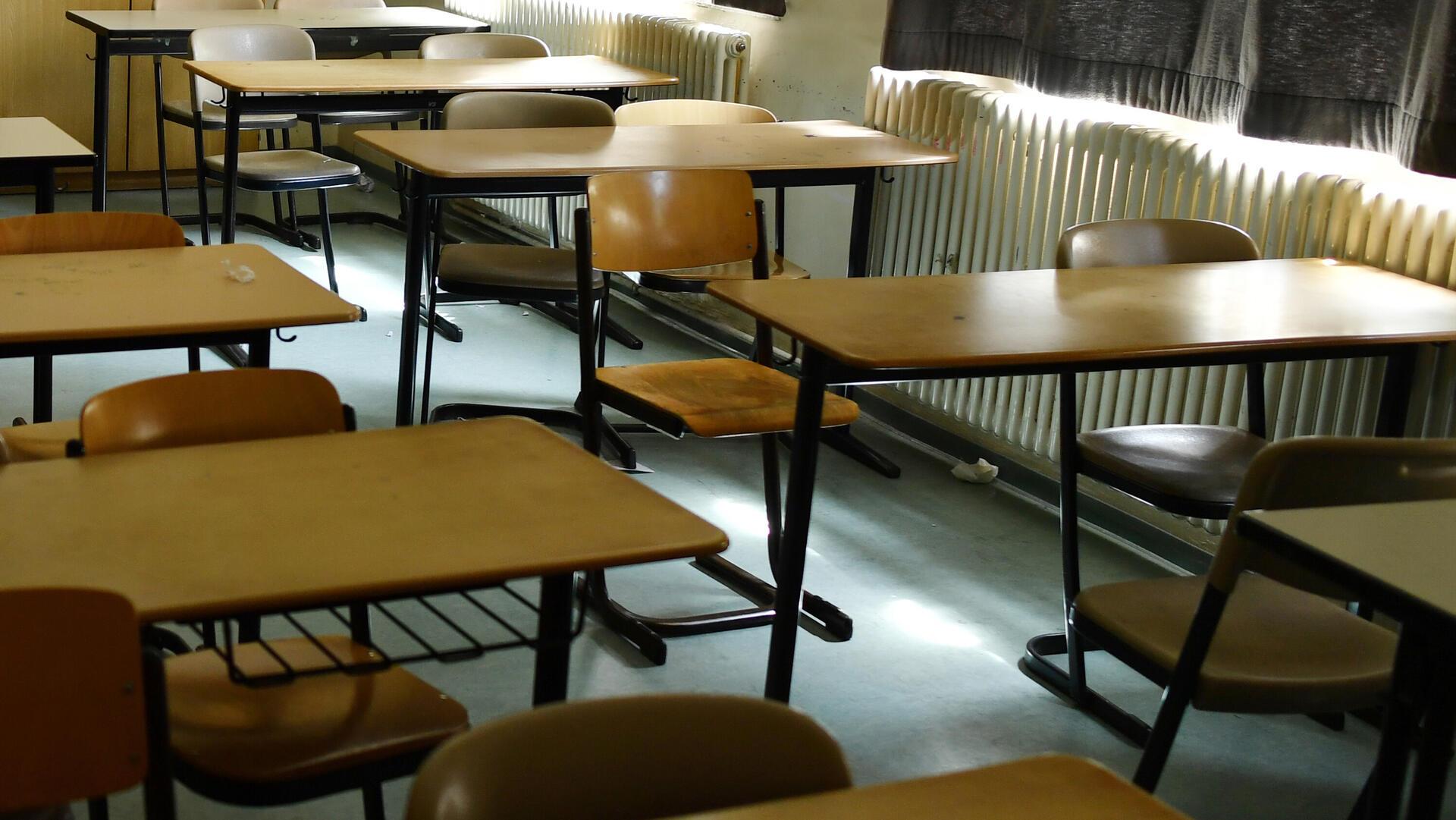 Leeres Klassenzimmer mit mehreren Schultischen und Stühlen, durch ein Fenster fällt Licht auf den Boden.