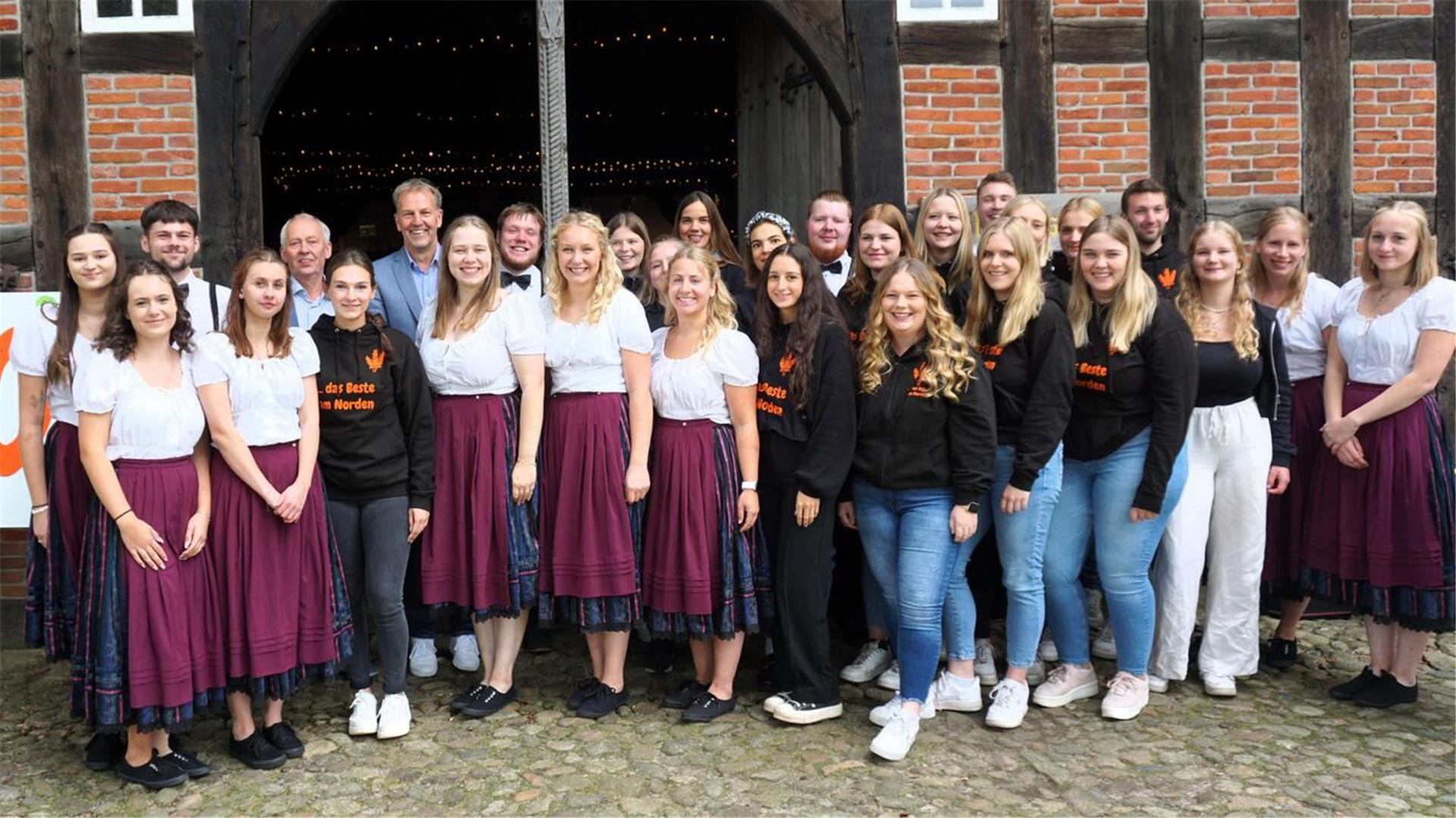 **ALT-Text:**
Eine große Gruppe junger Frauen und Männer, viele in Tracht oder mit schwarzen Landjugend-Pullovern, steht lächelnd vor einem historischen Fachwerkhaus mit offener Tür.