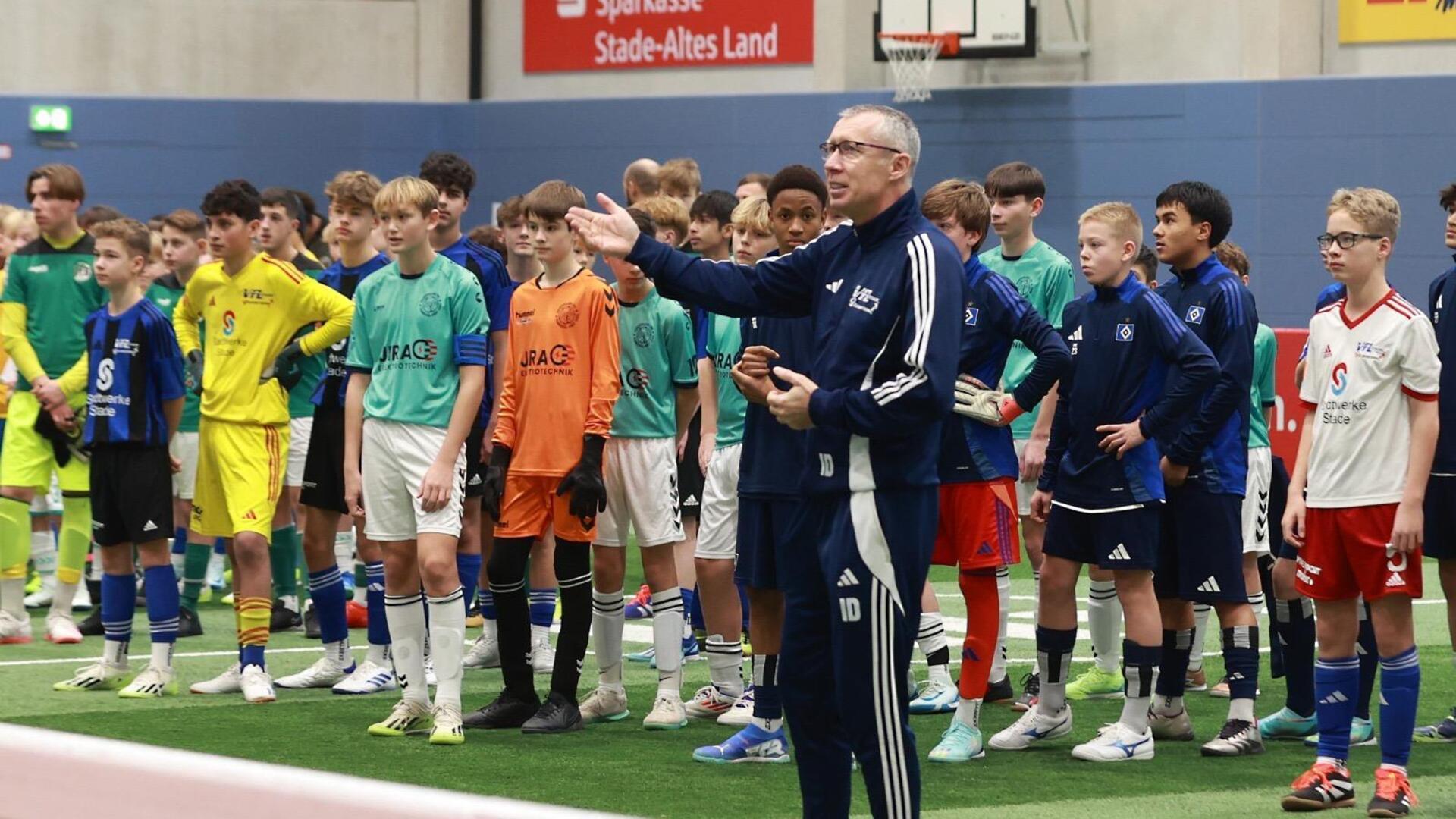 Ein Mann in einem blauen Trainingsanzug steht in einer Halle vor vielen Jungen in Sporttrikots.