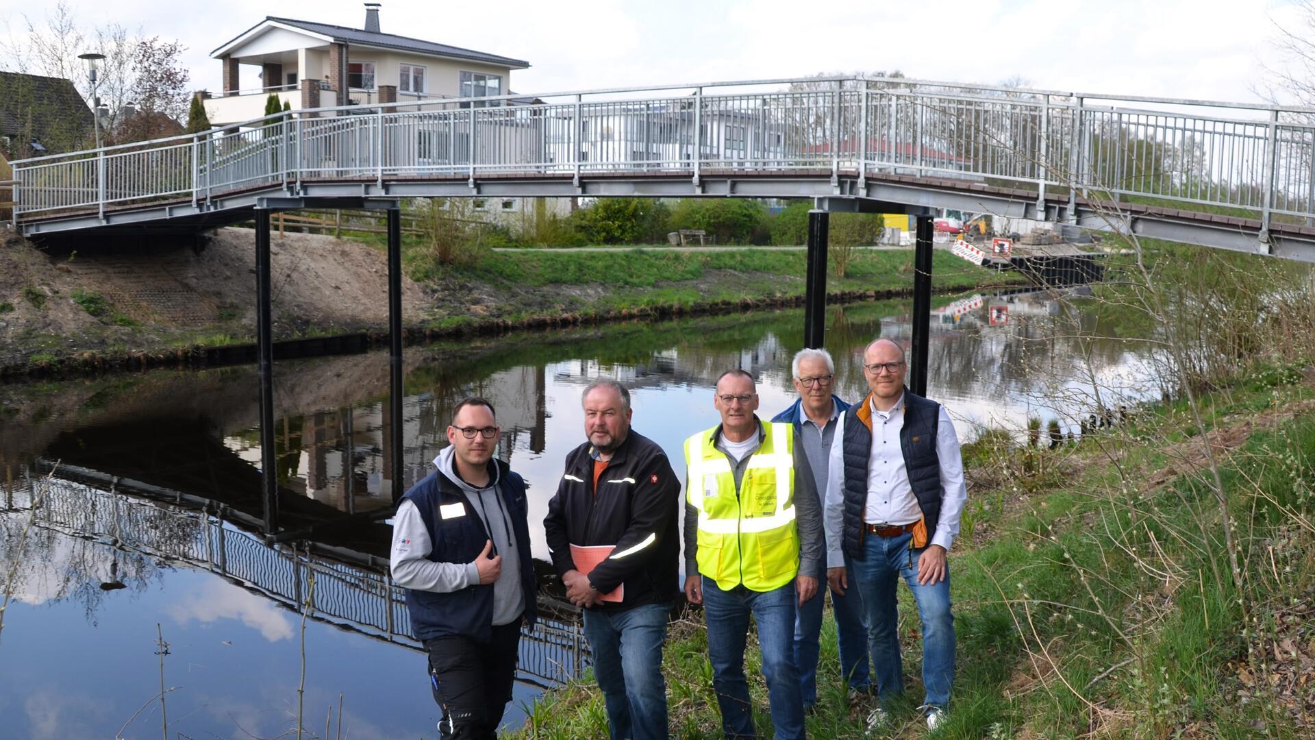 Fünf Männer stehen vor einer neu errichteten Fußgängerbrücke über den Hadelner Kanal in Geestland. Hinter ihnen verläuft die Brücke mit Metallgeländer über das Wasser. 