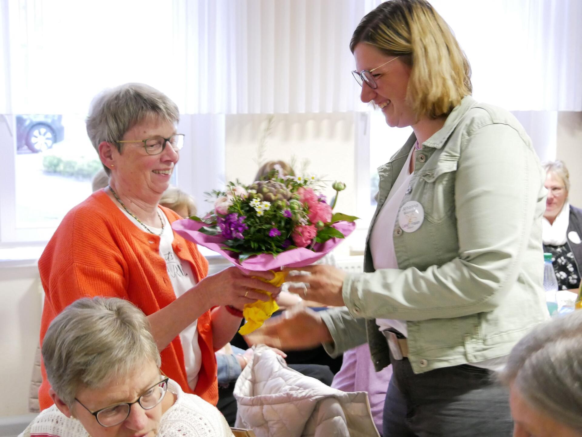 Eine Frau in oranger Strickjacke bekommt einen Blumenstrauß überreicht.