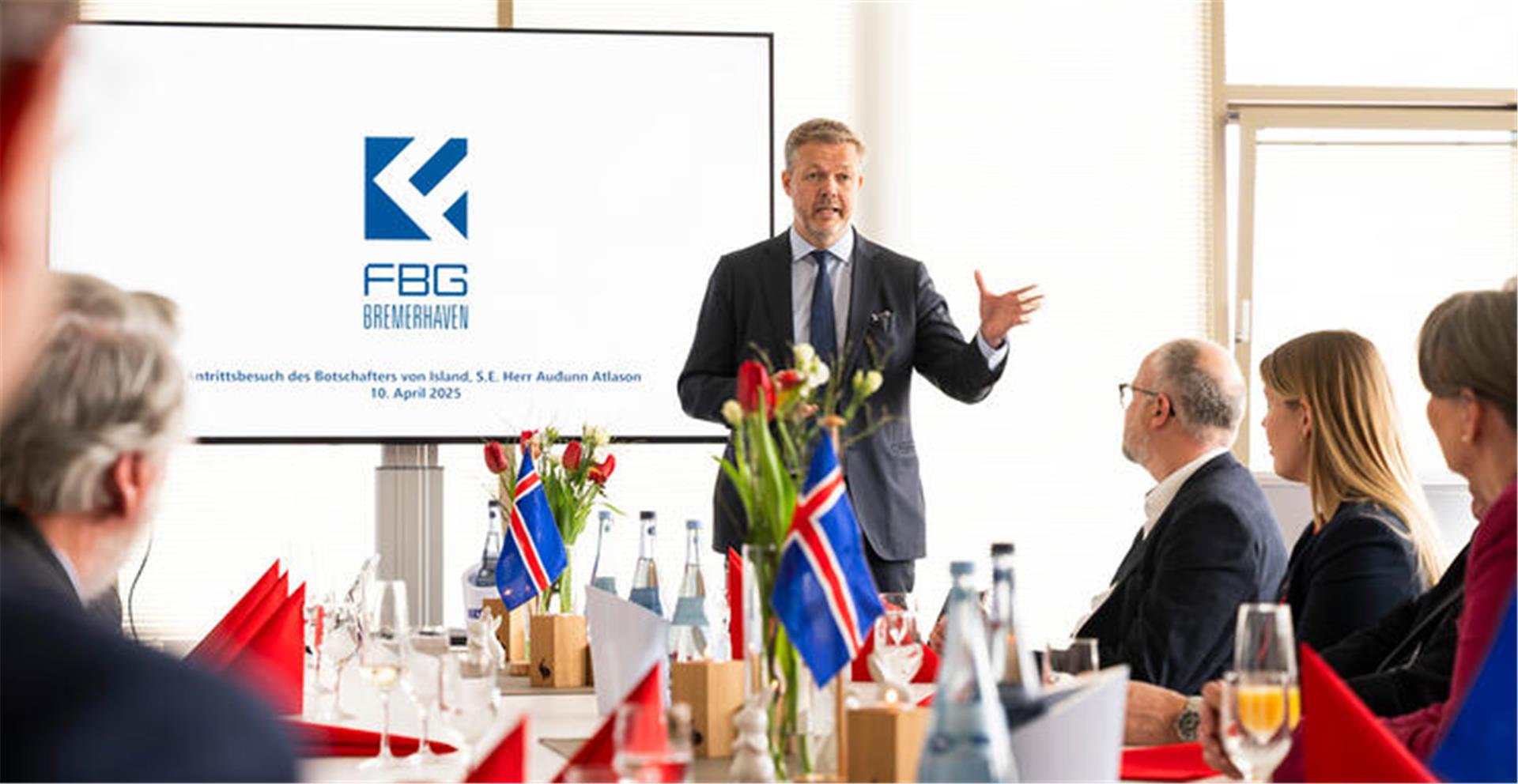 Ein Mann spricht stehend vor mehreren sitzenden Menschen. Auf einem großen BIldschirm hinter ihm ist das Wort "FBG" und das blau-weiße Logo des Unternehmens zu lesen. Island und Bremerhaven verbindet eine lange Partnerschaft in der Fischwirtschaft – Botschafter Auðunn Atlason würdigte beim Besuch des Fischereihafens die enge und verlässliche Zusammenarbeit. 