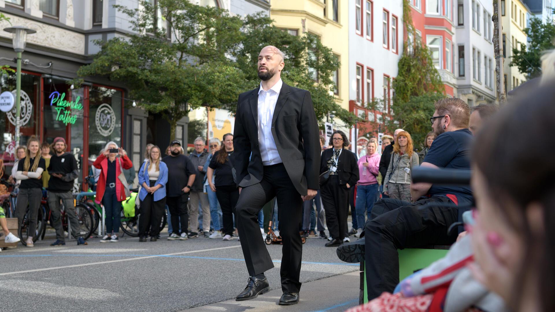 Ein Mann geht auf einer Stra0e - scheinbar, beobachtet von vielen Menschen. Ist das noch die Straße oder doch ein Turnierplatz? Paul Hess verwandelt sich in ein Pferd, nicht in irgendeines, sondern in den Wunderhengst Totila, der mehrere Goldmedaillen gewann.