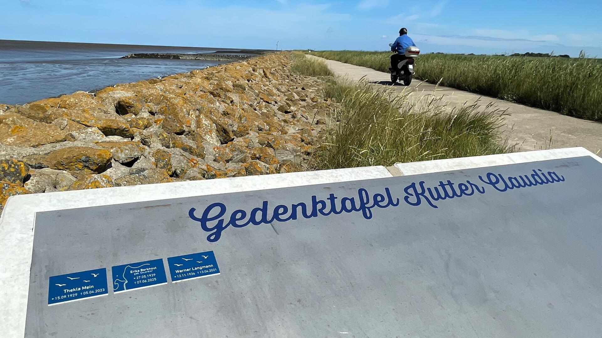 Die "Gedenktafel Kutter Claudia" an einer felsigen Küste. Im Hintergrund verläuft ein Strandweg, auf dem ein Mopdefahrer fährt.