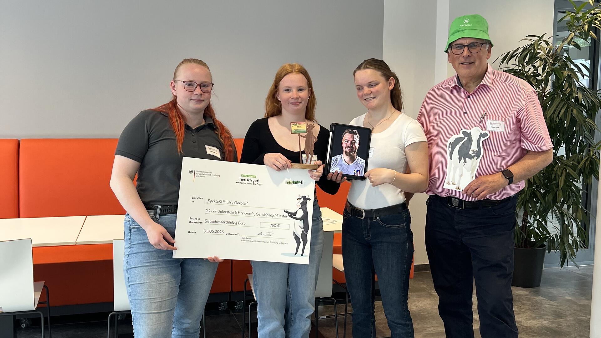 Drei Schülerinnen des GenoKollegs Münster stehen mit Lehrer Klaus Feils in einem modernen Raum. Sie halten einen Scheck über 750 Euro, einen Pokal, ein Tablet mit einem Porträt und eine Kuhfigur – als Auszeichnung für den dritten Platz beim Wettbewerb „Echt kuh-l! 2025“.