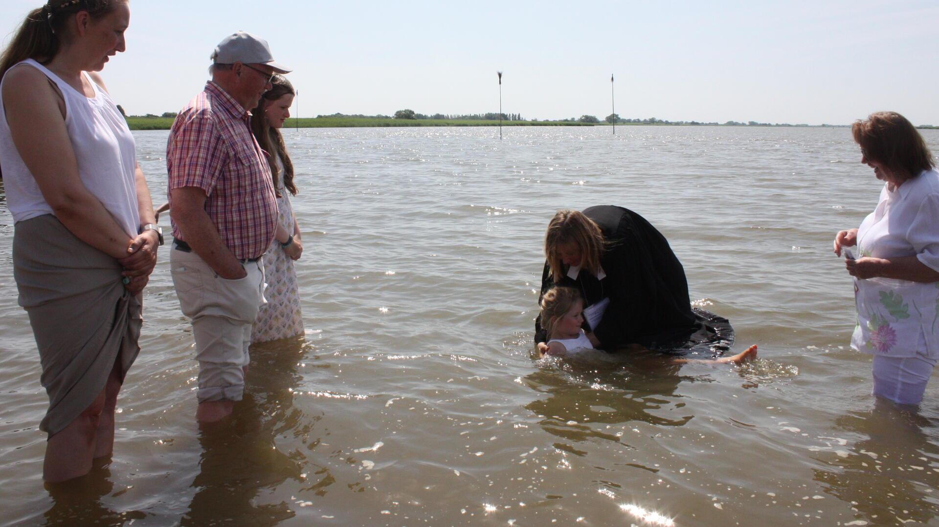 Pastorin tauft ein Mädchen in der Weser während eines Taufgottesdienstes; um sie herum stehen Familienangehörige und weitere Teilnehmende im Wasser.