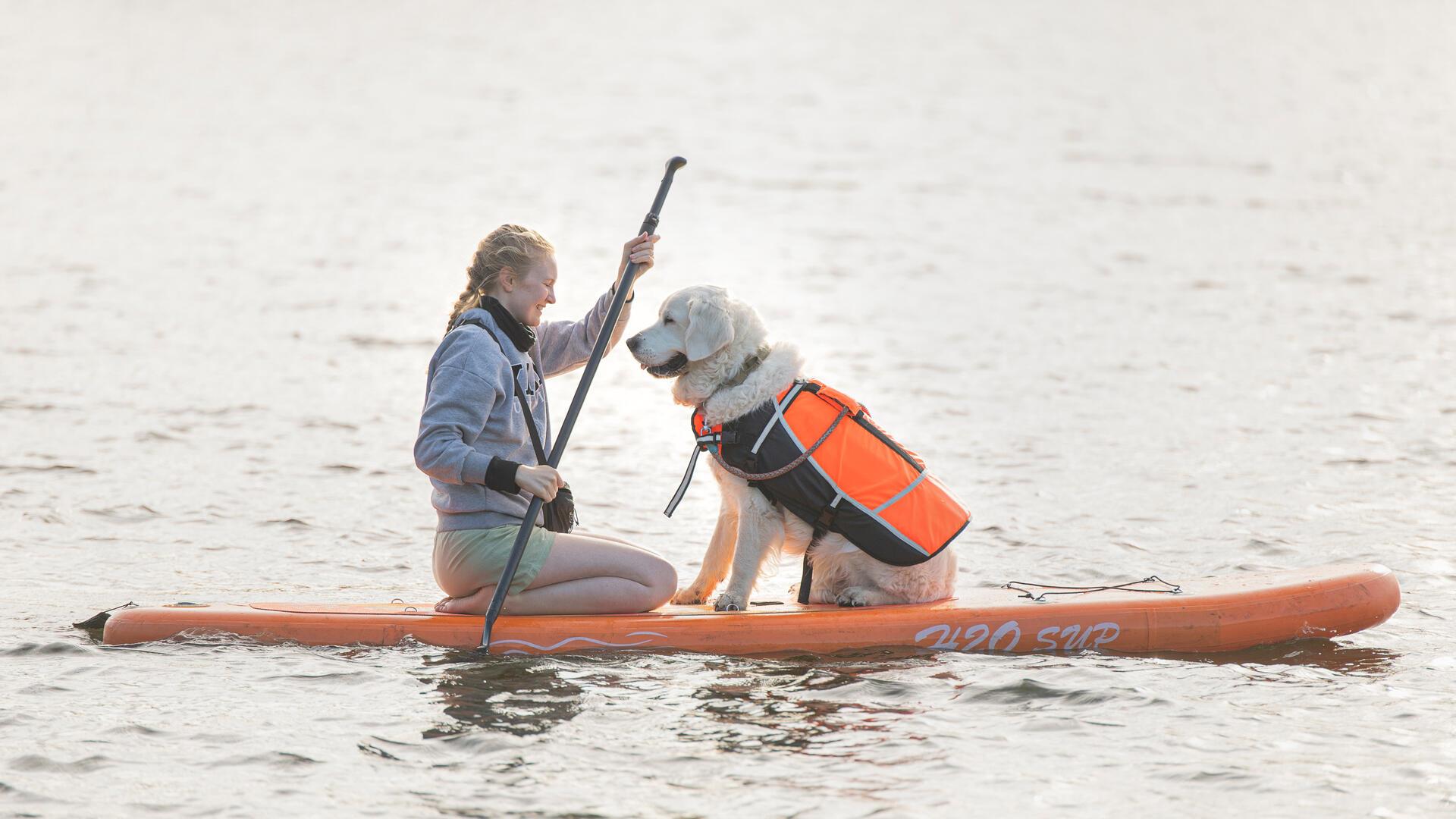 Eine Frau und ein Hund sitzen auf einem Stand-up-Paddleboard und schauen sich an. Die Frau hält ein Paddel in den Händen.