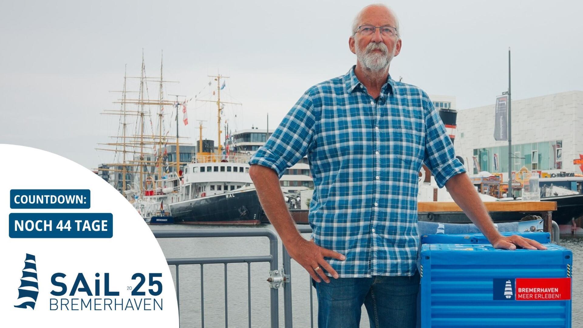 Ein Mann mit kariertem Hemd steht am Hafen von Bremerhaven vor Segelschiffen. Im Bild ist ein Countdown zur Sail 2025 mit „Noch 44 Tage“ eingeblendet.