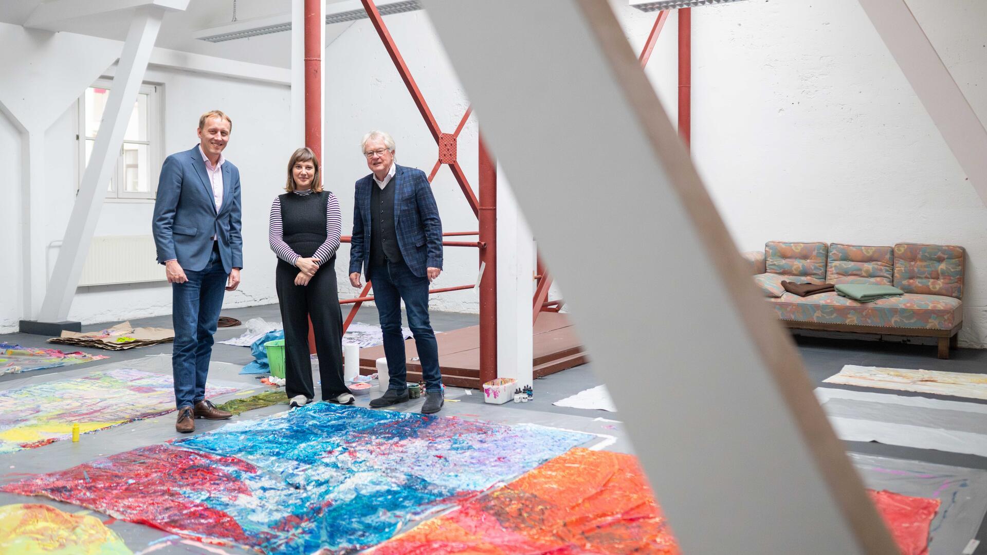 Drei Personen stehen in einem großzügigen, hellen Atelierraum zwischen großformatigen, farbenfrohen Kunstwerken, die auf dem Boden ausgebreitet sind. Im Hintergrund sind rote Stahlträger, weiße Wände und ein Sofa zu sehen.