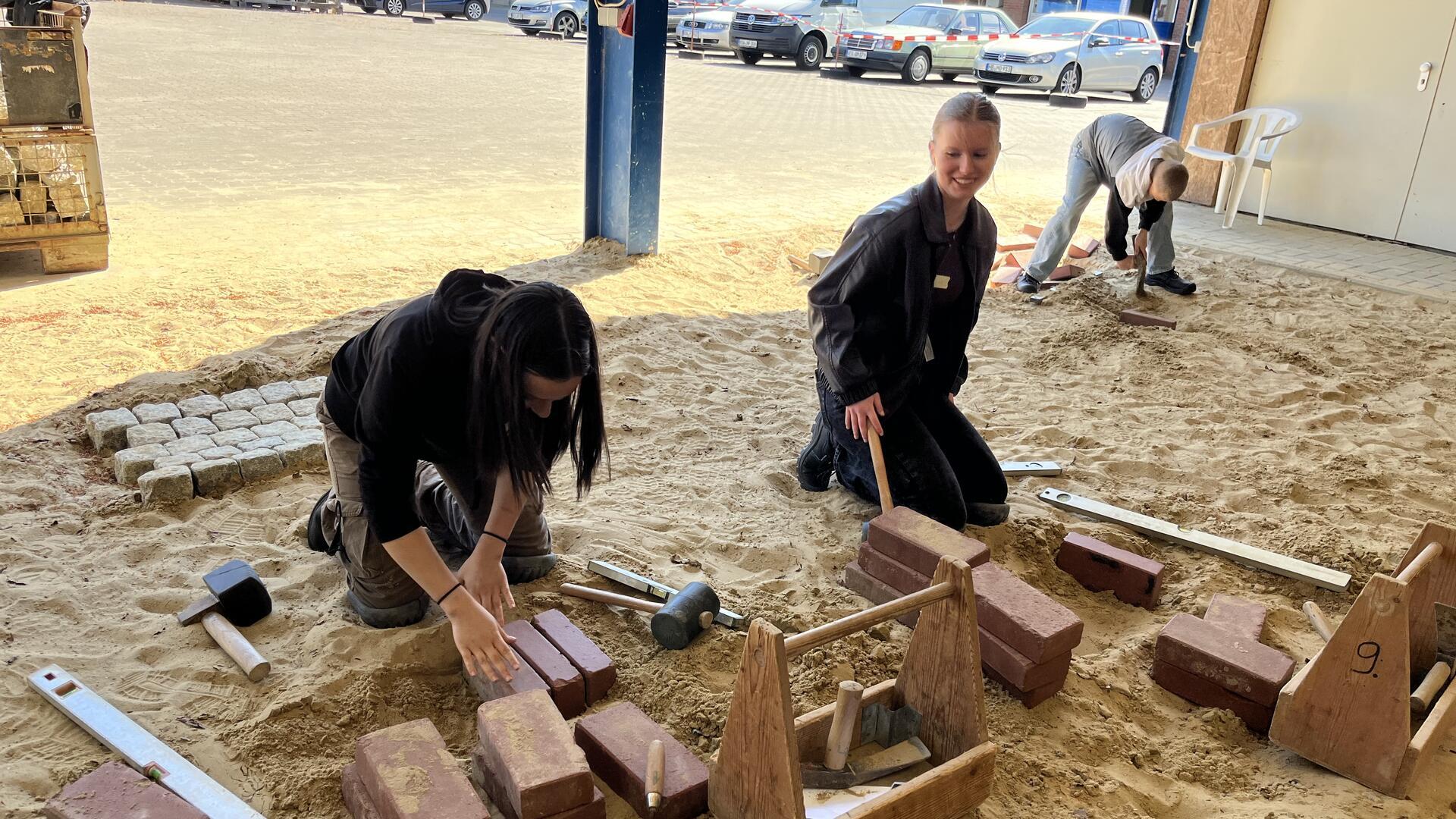 Zwei Mädchen knien in Sand und bearbeiten Pflastersteine.