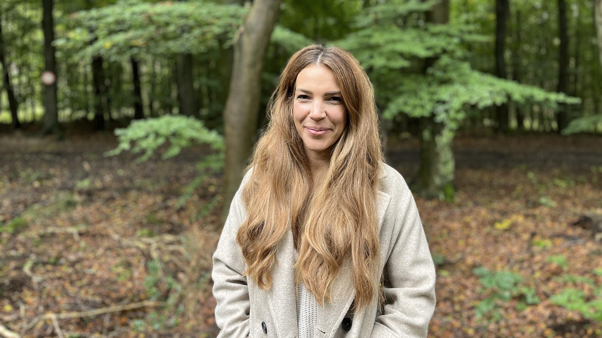 Eine Frau mit langen, braunen Haaren und einem beigefarbenen Mantel steht in einem Wald.