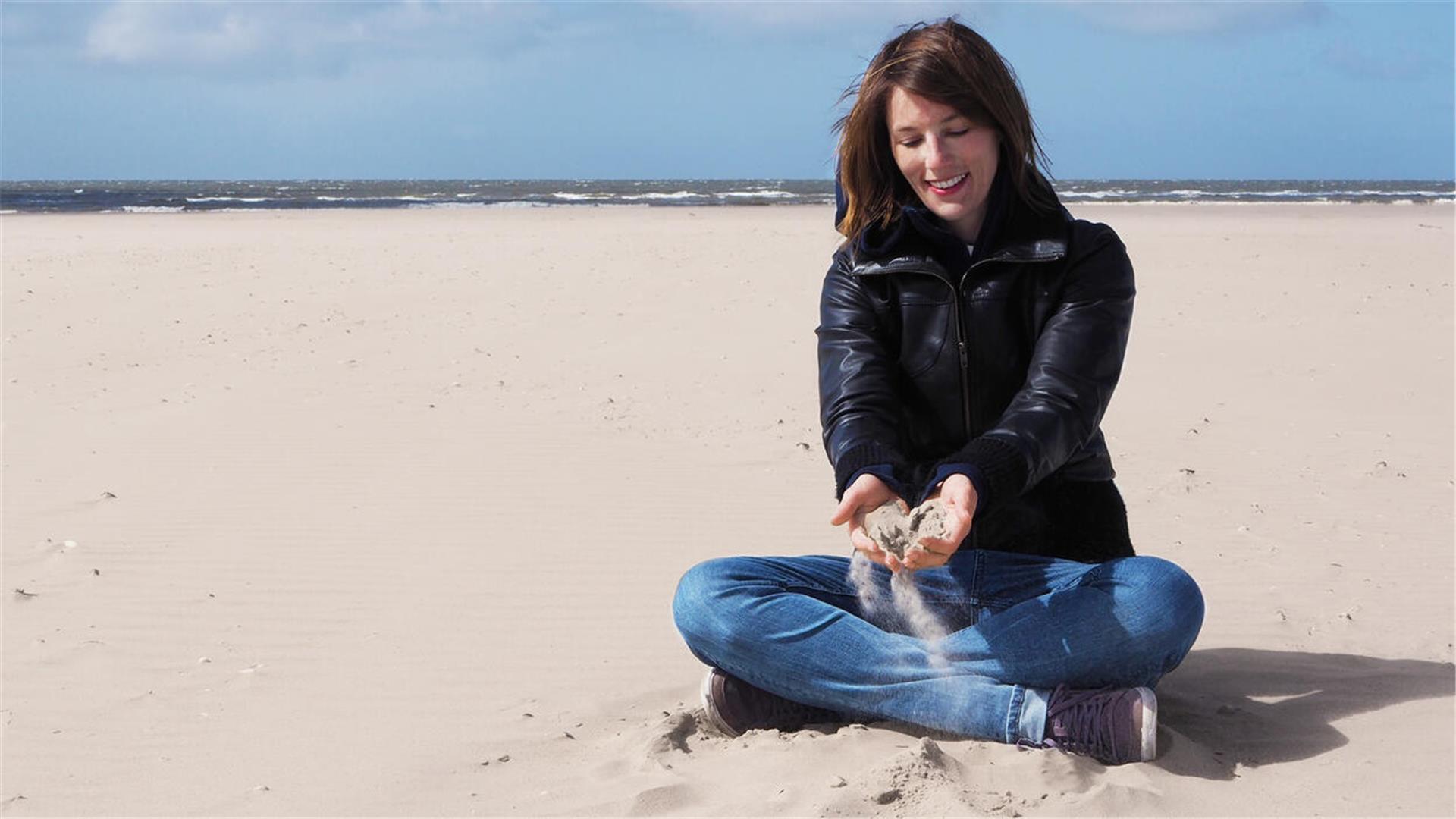 Frau sitzt im Sand am Strand und hält Sand in den Händen