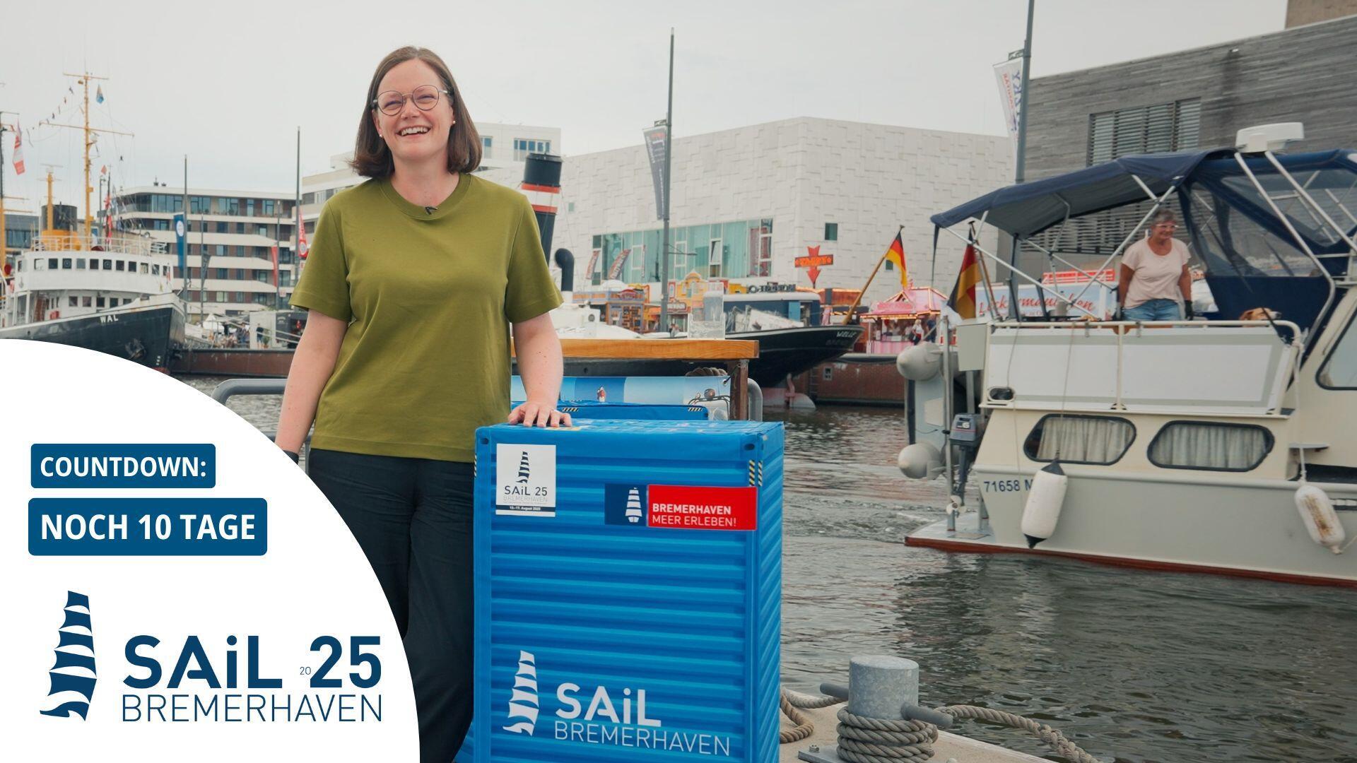 Eine Frau steht lächelnd vor maritimer Kulisse in Bremerhaven neben einem blauen Container mit dem Logo der Sail 2025. Im Hintergrund sind Schiffe, Hafenanlagen und ein Festbereich zu sehen.