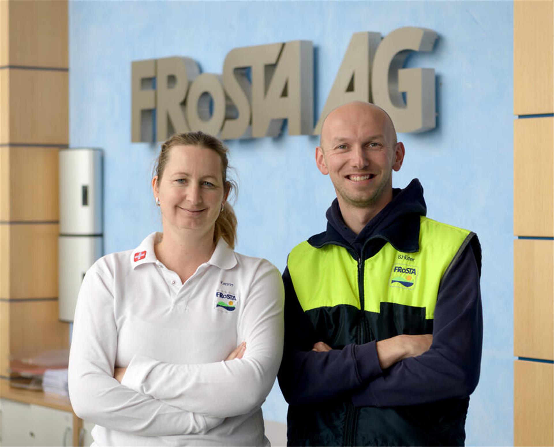 Eine Frau und ein Mann mittleren Alters lächeln in die Kamera vor dem Schriftzug Frosta AG im Hintergrund.  