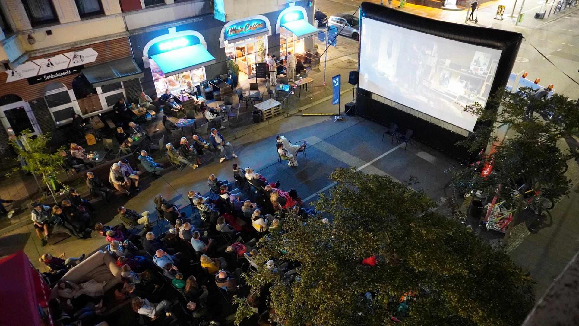 Zahlreiche Menschen sitzen abends auf Klappstühlen und Sofas vor einer großen Leinwand bei einem Open-Air-Kino in der Alten Bürger in Bremerhaven.