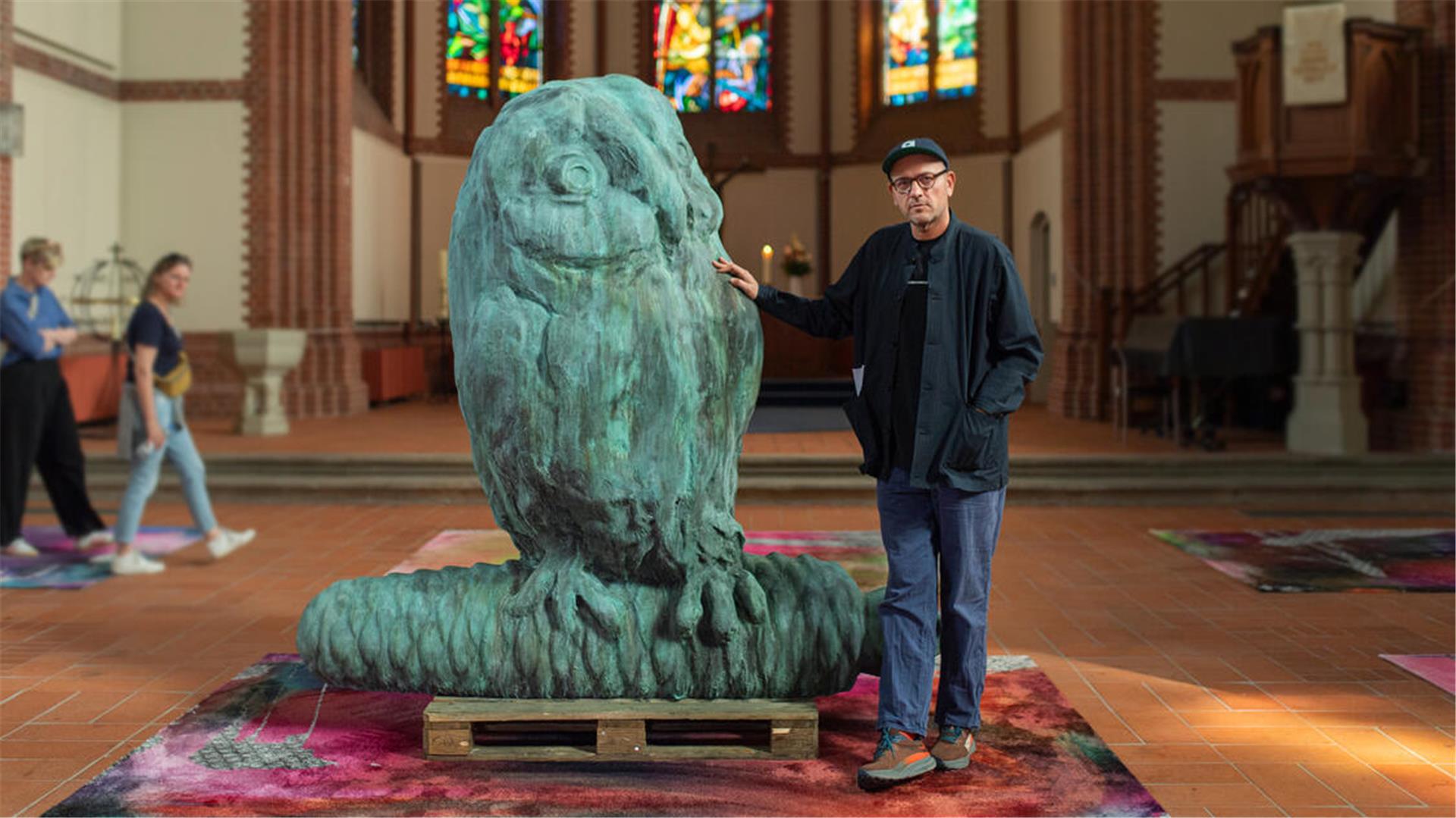 In einer Kirche steht eine mehr als zwei Meter große Eule aus Bronze, daneben steht ein Mann. Auf dem Boden liegen bunte Teppiche.
