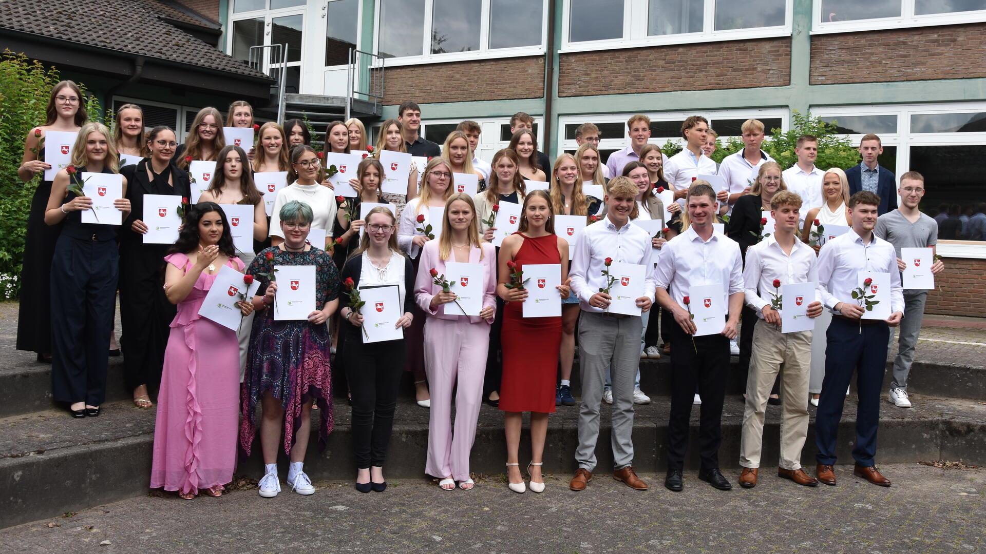 41 Schülerinnen und Schüler stehen in festlicher Kleidung auf einer Treppe für ein Foto bereit und halten ihre Zeugnisse.