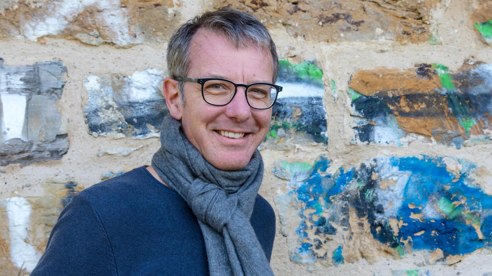 Ein Mann mit blauem Pullover steht vor einer Wand mit blauer Farbe.