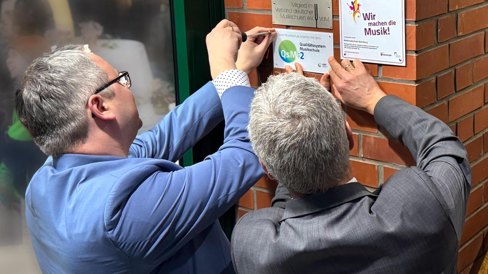 Landrat Marco Prietz (links) und Bürgermeister Torsten Oestmann bringen das Siegel am Gebäude der Kreismusikschule an.