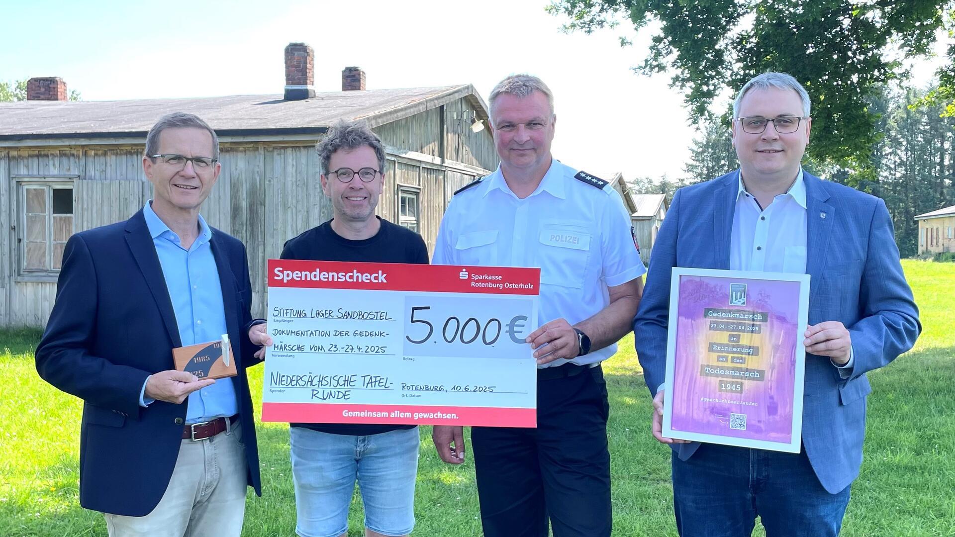 Vier Männer halten einen symbolischen Spendenscheck vor einem Gebäude in den Händen. Landrat Marco Prietz (rechts) und Stefan Kalt (links) überreichen den Spendenscheck an Michael Freitag-Parey (Zweiter von links) von der Gedenkstätte Lager Sandbostel und Michael Butt vom Polizeikommissariat Zeven. Die 5.000 Euro der Niedersächsischen Tafelrunde fließen in eine Videodokumentation zum Gedenkmarsch „Steps to remember“.