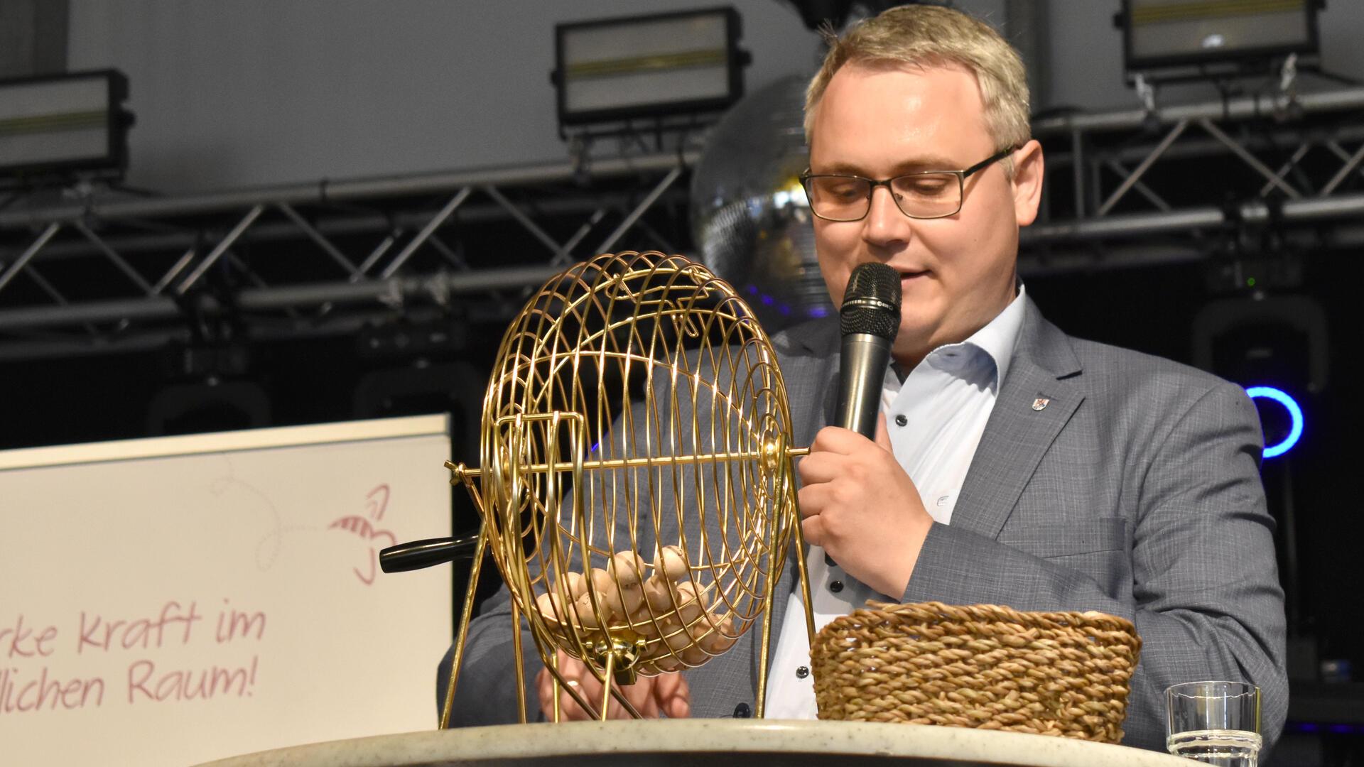Ein Mann hat einen runden Korb aus Metall vor sich auf einem Tisch stehen und spricht in ein Mikrofon.