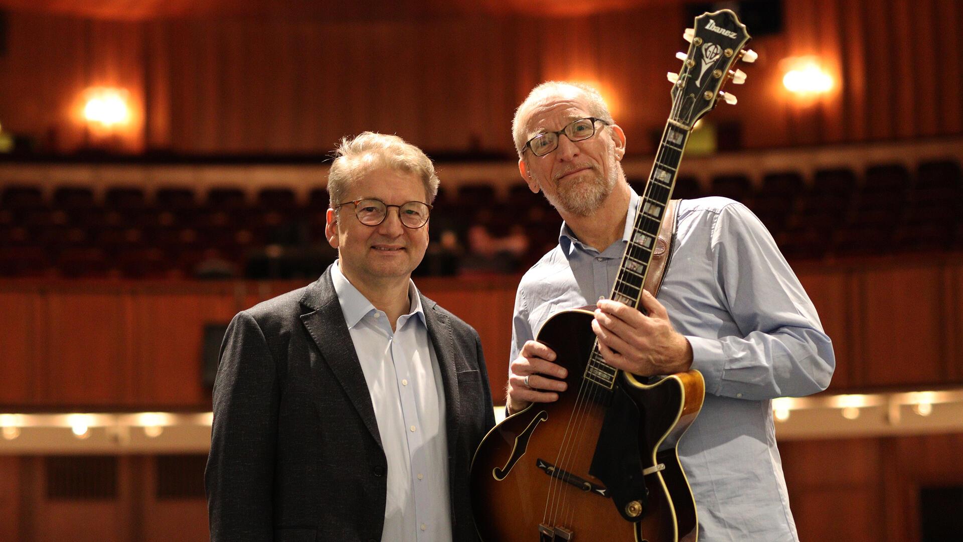 Zwei Männer stehen im Zuschauerraum eines Theaters. Der Mann rechts hält eine Jazzgitarre, beide lächeln in die Kamera. Im Hintergrund sind leere Sitzreihen und warmes Licht zu sehen.