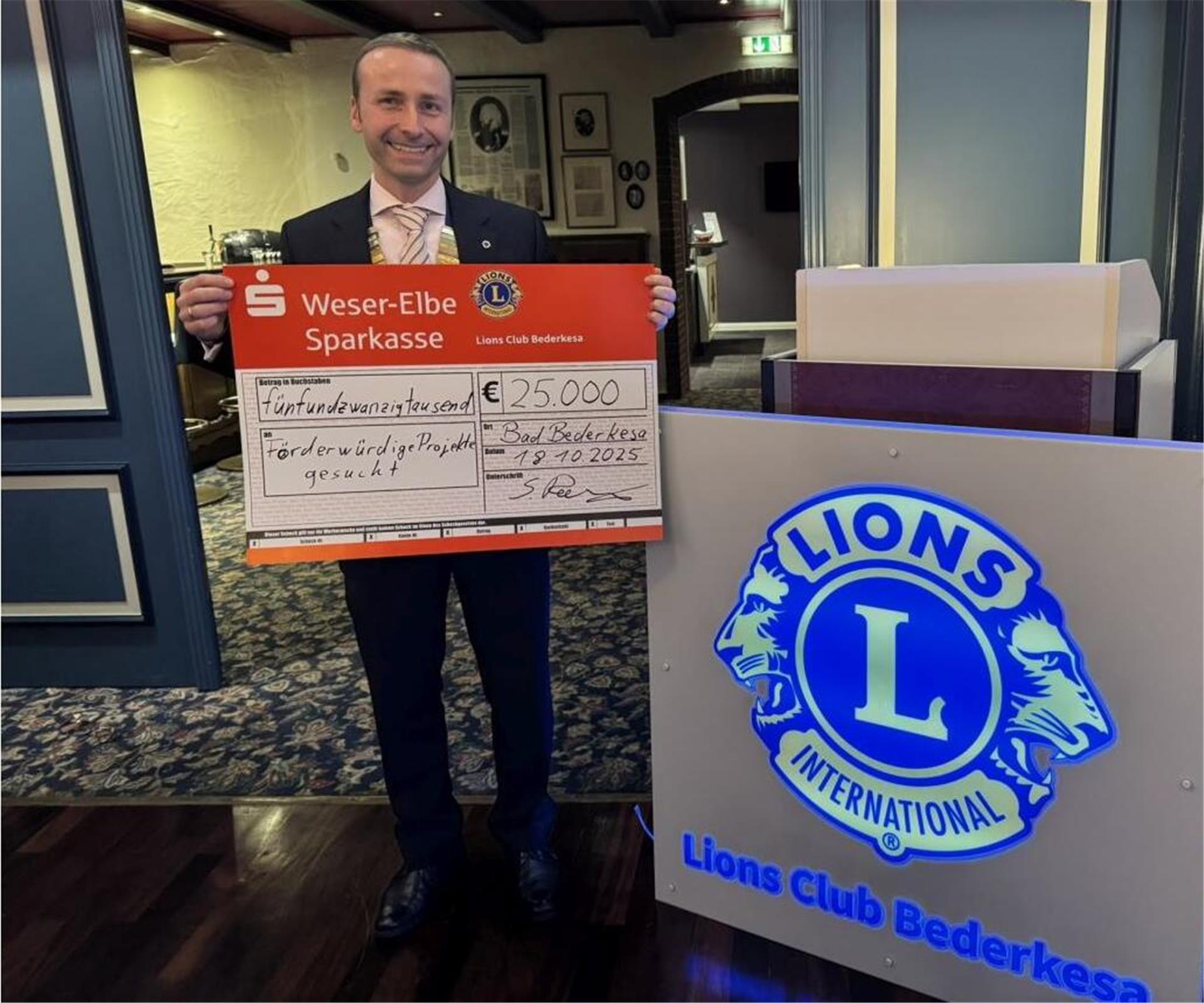 Ein Mann im Anzug hält einen großen symbolischen Spendenscheck über 25.000 Euro in den Händen. Neben ihm steht ein beleuchtetes Schild mit dem Logo des Lions Club Bederkesa.
