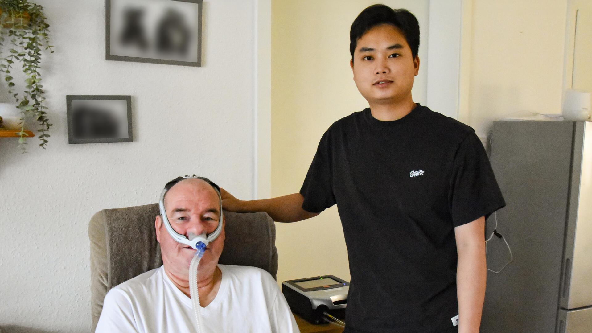 Ein älterer Mann mit Maskenbeatmung sitzt lächelnd auf einem Sessel, neben ihm steht ein junger Mann.
