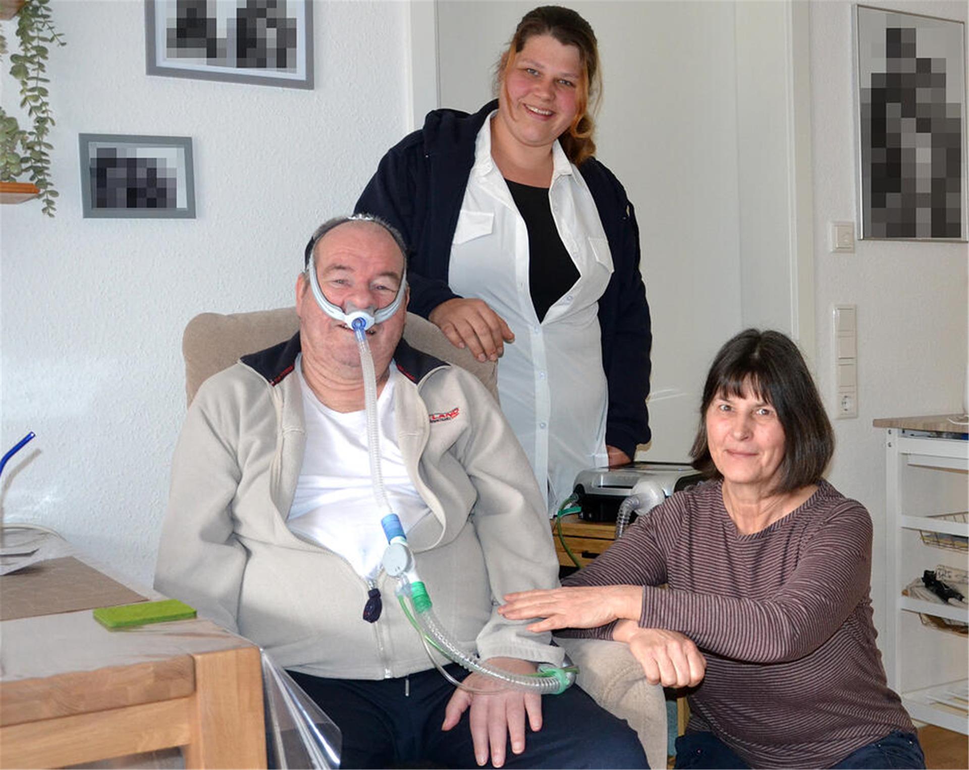 Ein Mann mit Beatmungsmaske sitzt mit wachem Blick in einem Sessel, hinter und neben ihm sind zwei Frauen, im Hintergrund ist das Beatmungsgerät erkennbar.