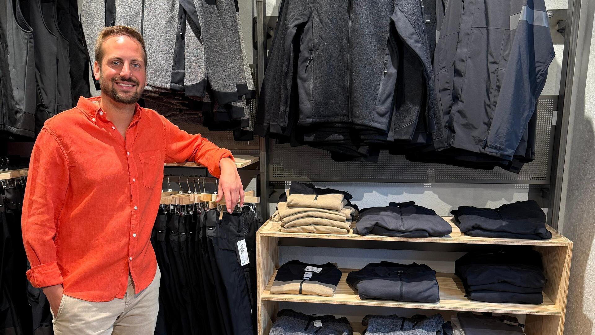 Ein junger Mann im orangefarbenen Hemd steht neben einem Regal, in dem zusammengefaltete Kleidung liegt, hinter ihm hängen Jacken auf Kleiderbügeln.