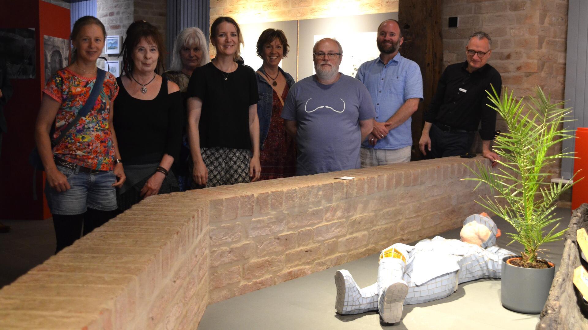 Gruppe von neun Personen steht in einem Ausstellungsraum hinter einer niedrigen Backsteinmauer. Im Vordergrund liegt eine Figur in Mosaikoptik neben einer Topfpflanze.